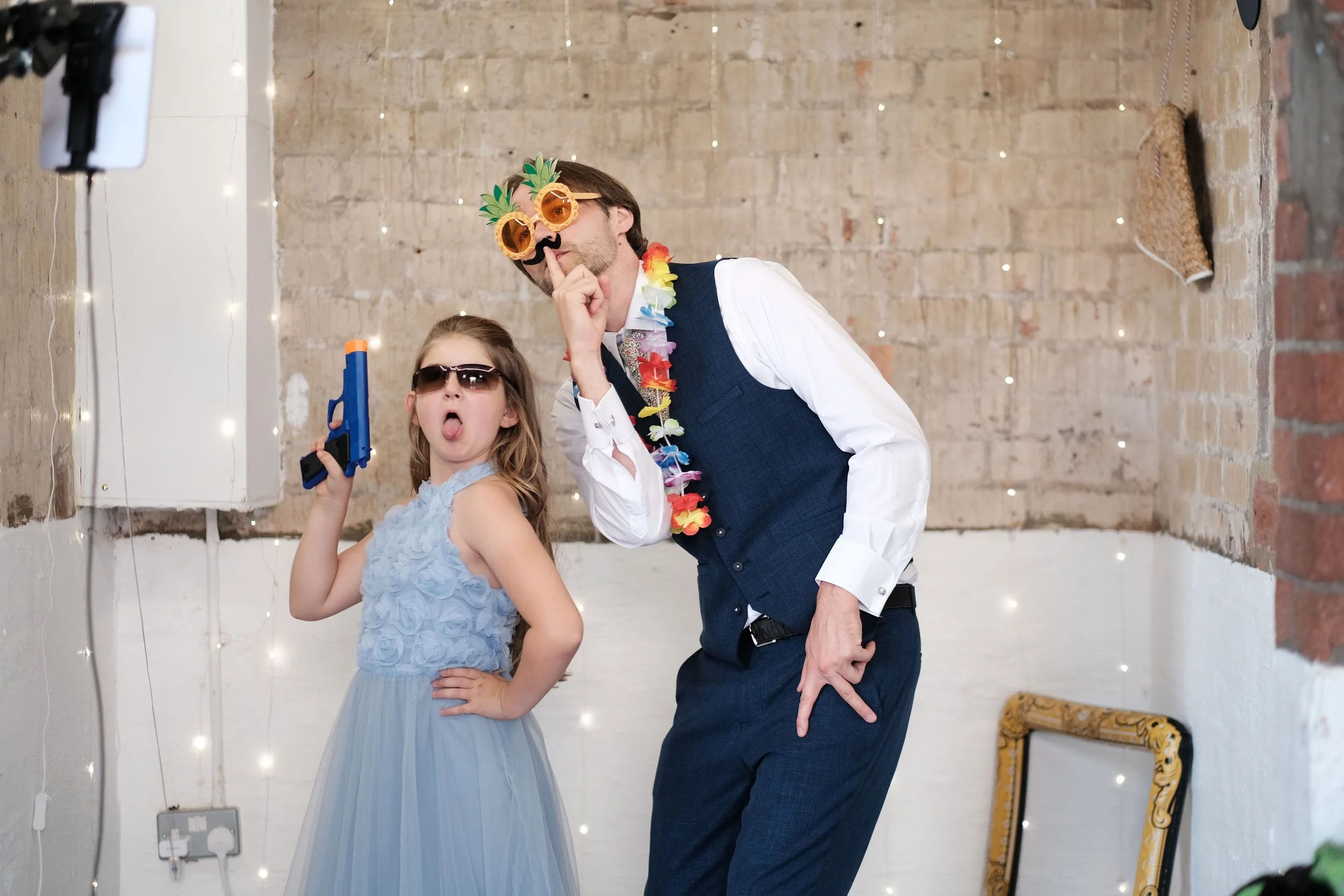 Man and girl in costumes posing at party, with man wearing pineapple sunglasses and lei, girl with sunglasses and toy gun, in decorated room.