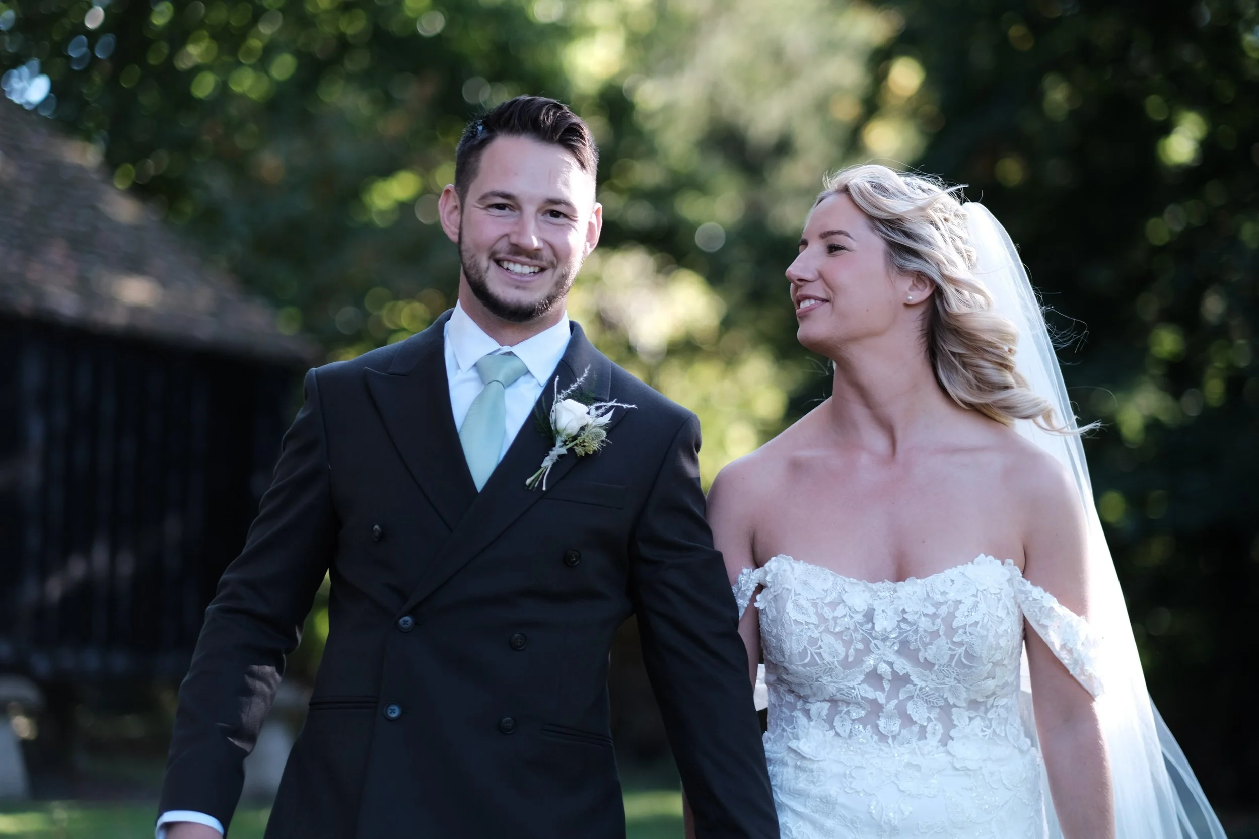 A newlywed couple walking outdoors, the groom wearing a black tuxedo with a light-colored tie and boutonniere, and the bride in an off-the-shoulder lace wedding dress with a veil.