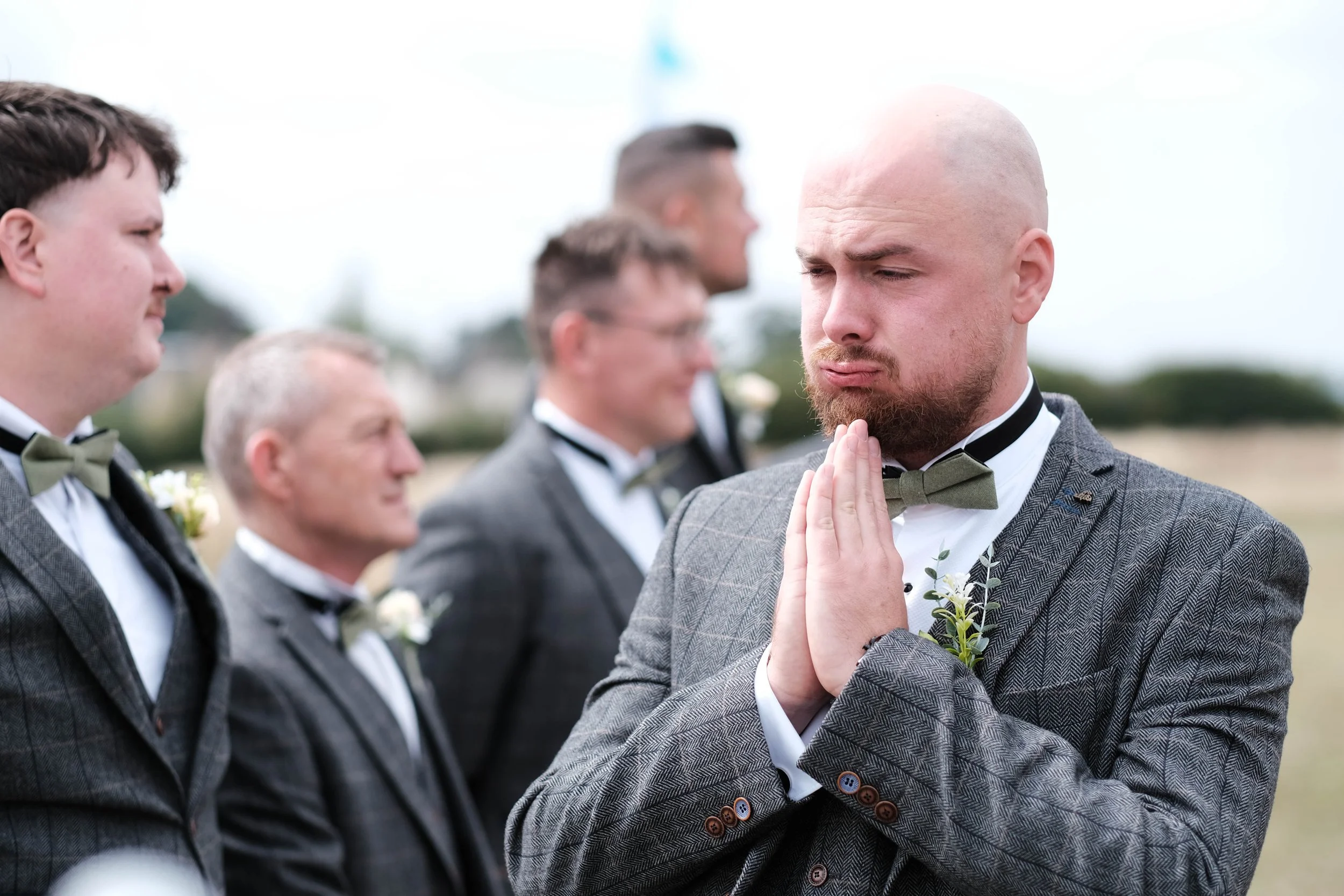 A man with a shaved head and beard in a gray suit praying with hands clasped at an outdoor wedding, with four other men in suits standing in line behind him.