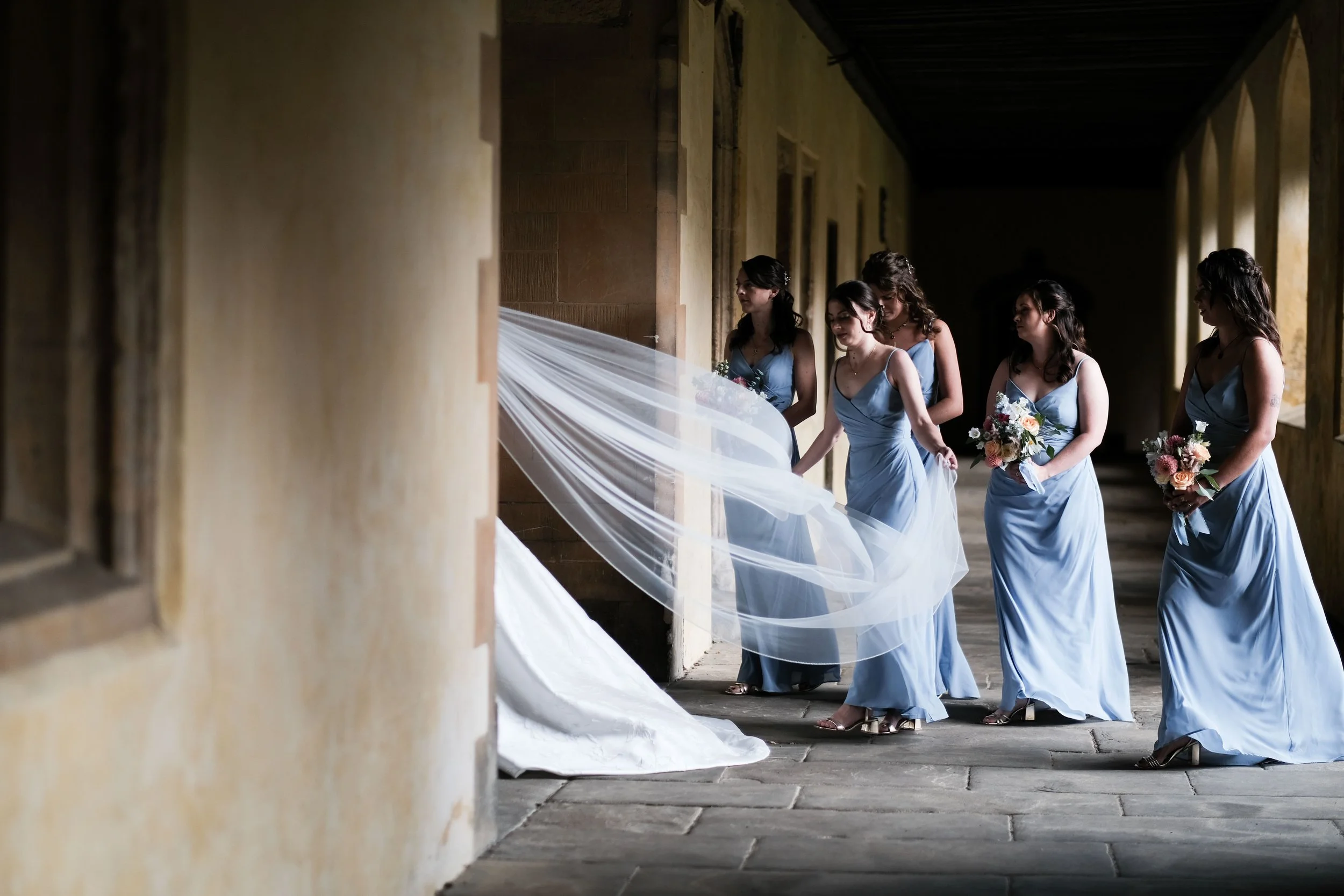 Five women in matching blue bridesmaid dresses standing in a corridor, one holding a bouquet, preparing for a wedding.