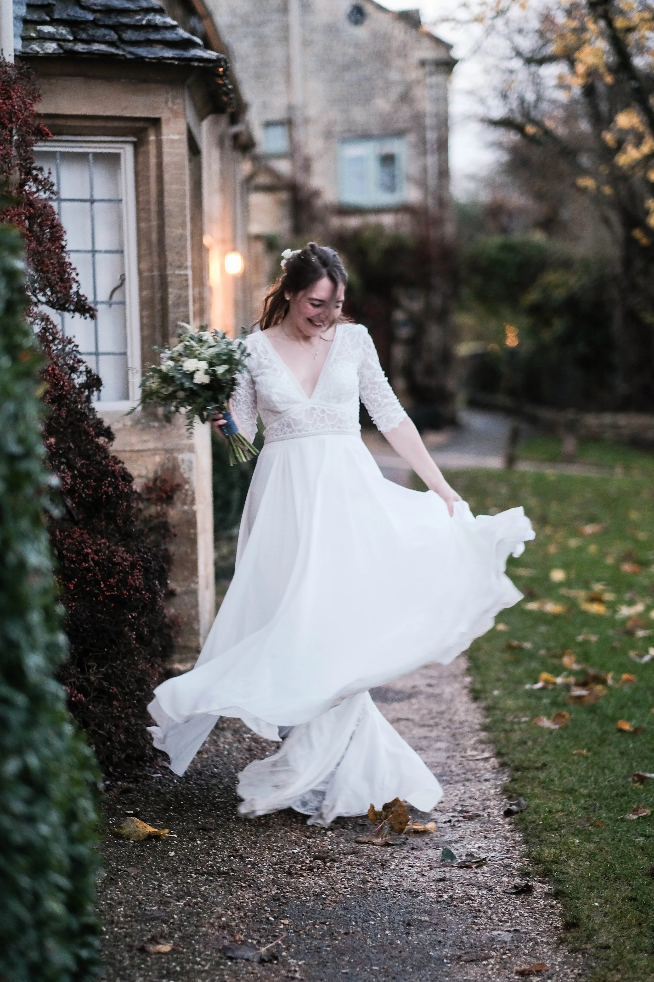 A bride in a white wedding dress holding a bouquet, smiling and twirling outside near a stone house and trees with fallen leaves.