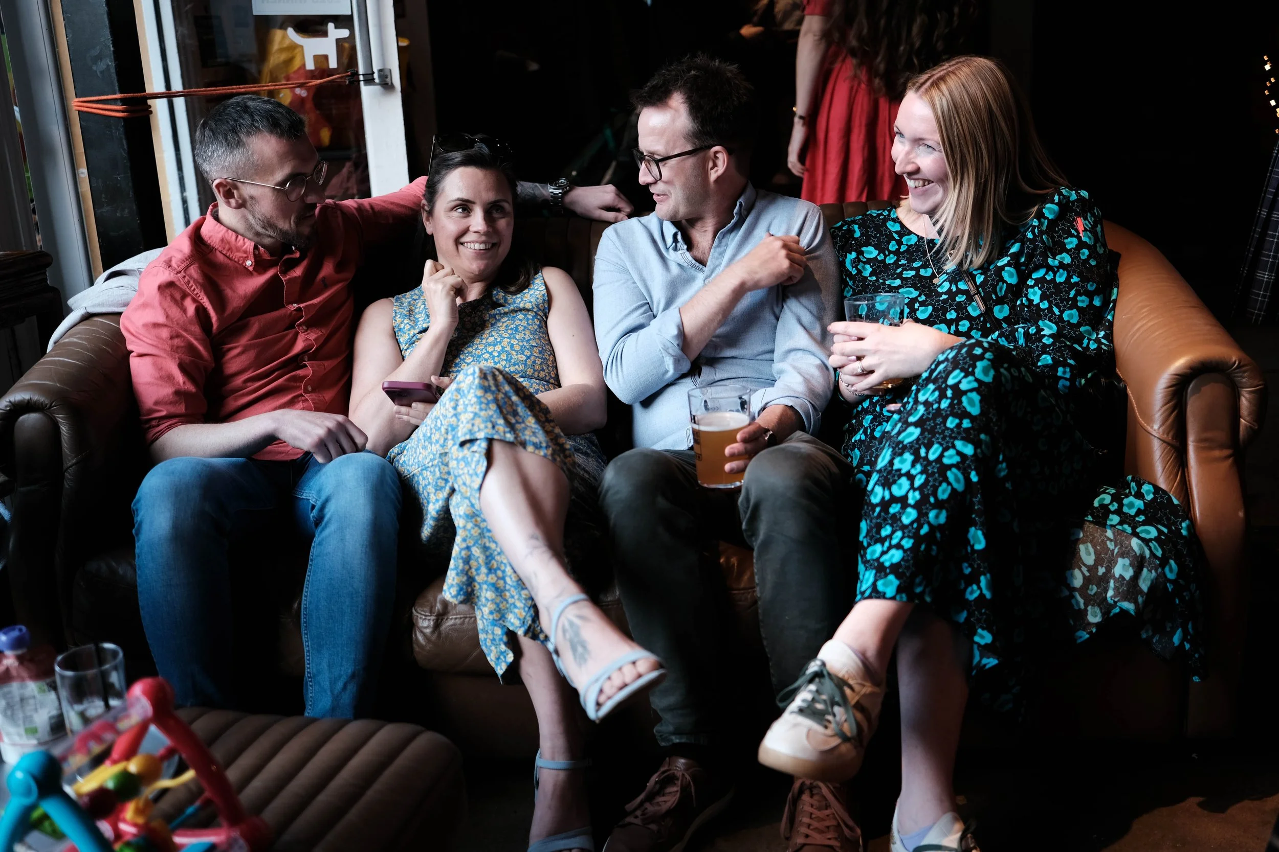 Four friends sitting closely on a leather couch in a living room, laughing and chatting. One man is wearing glasses and a light blue shirt, holding a beer. Two women are wearing patterned dresses, one holding a drink, and a man on the far left wearin