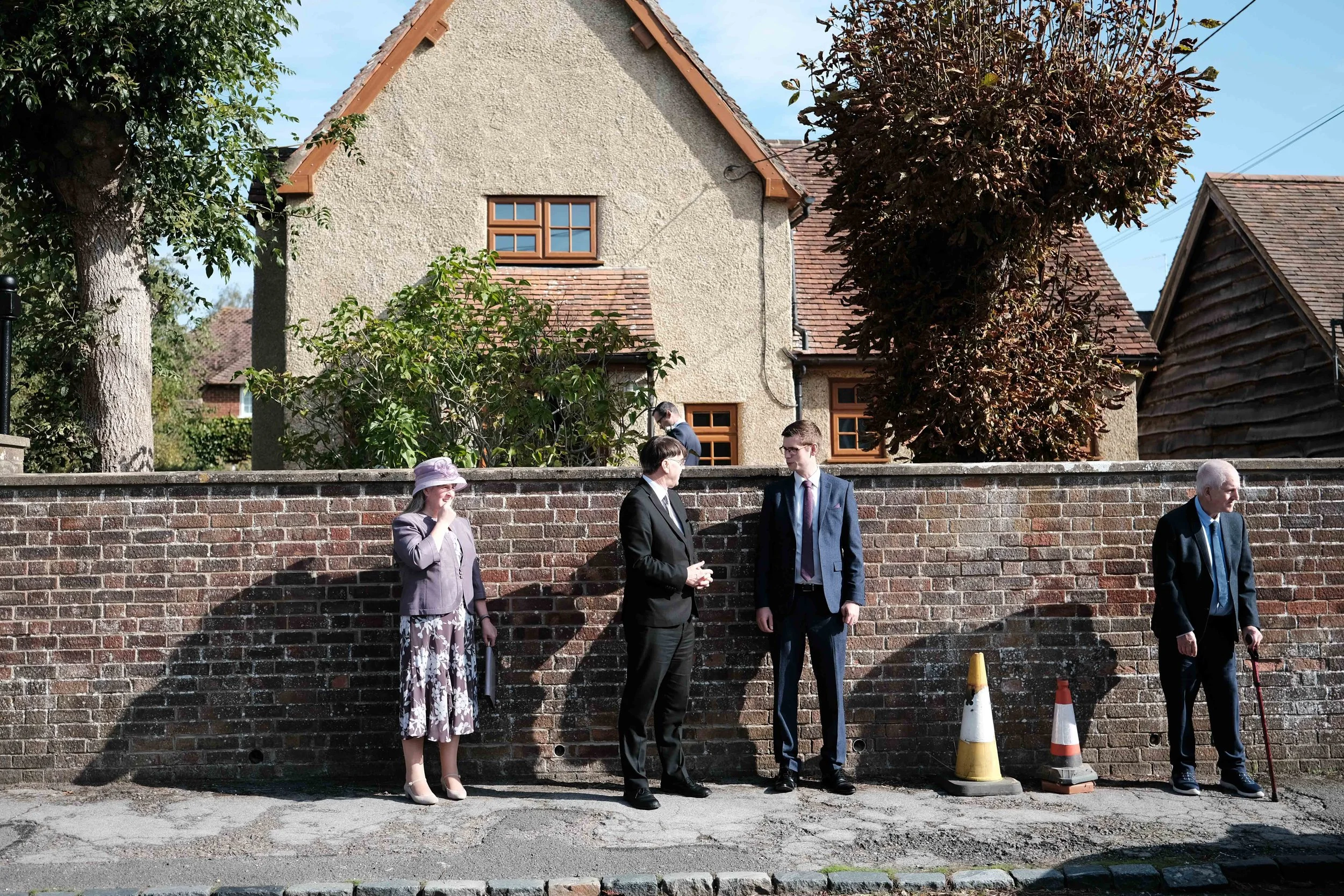 Four people standing in a line outdoors in front of a brick wall, engaging in conversation. A woman in a floral skirt and hat, and three men in suits, one elderly man with a cane. Two safety cones are on the ground near them. Behind the wall is a hou