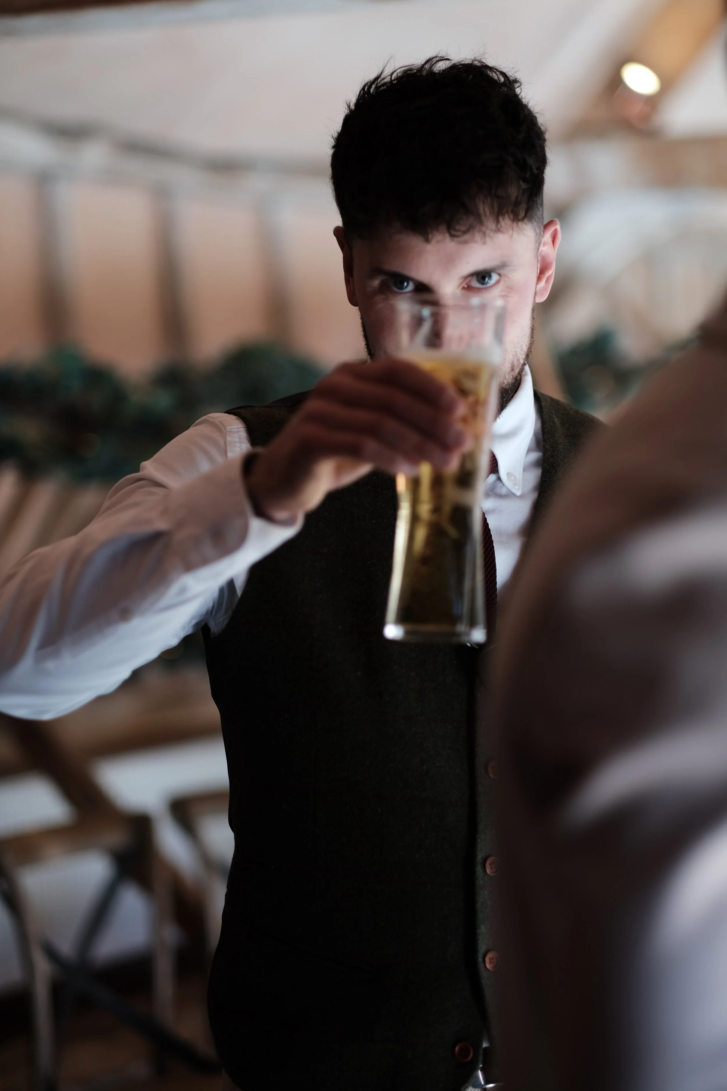 A man with dark hair and blue eyes holding a glass of beer in front of his face, looking directly at the camera.