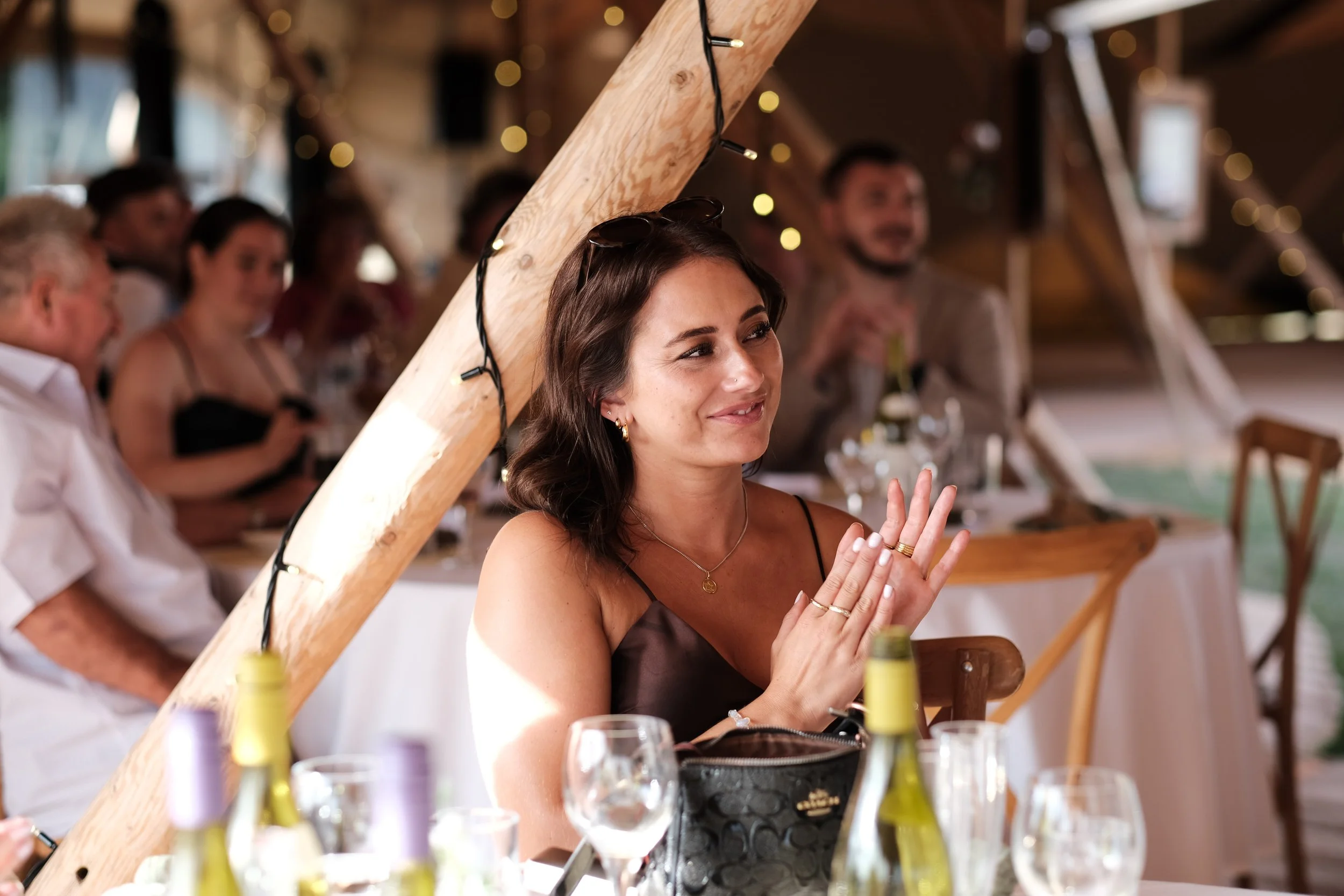 A woman with dark shoulder-length hair, wearing a black spaghetti strap dress and jewelry, smiling and clapping at a social event with people seated at tables in a decorated rustic venue.