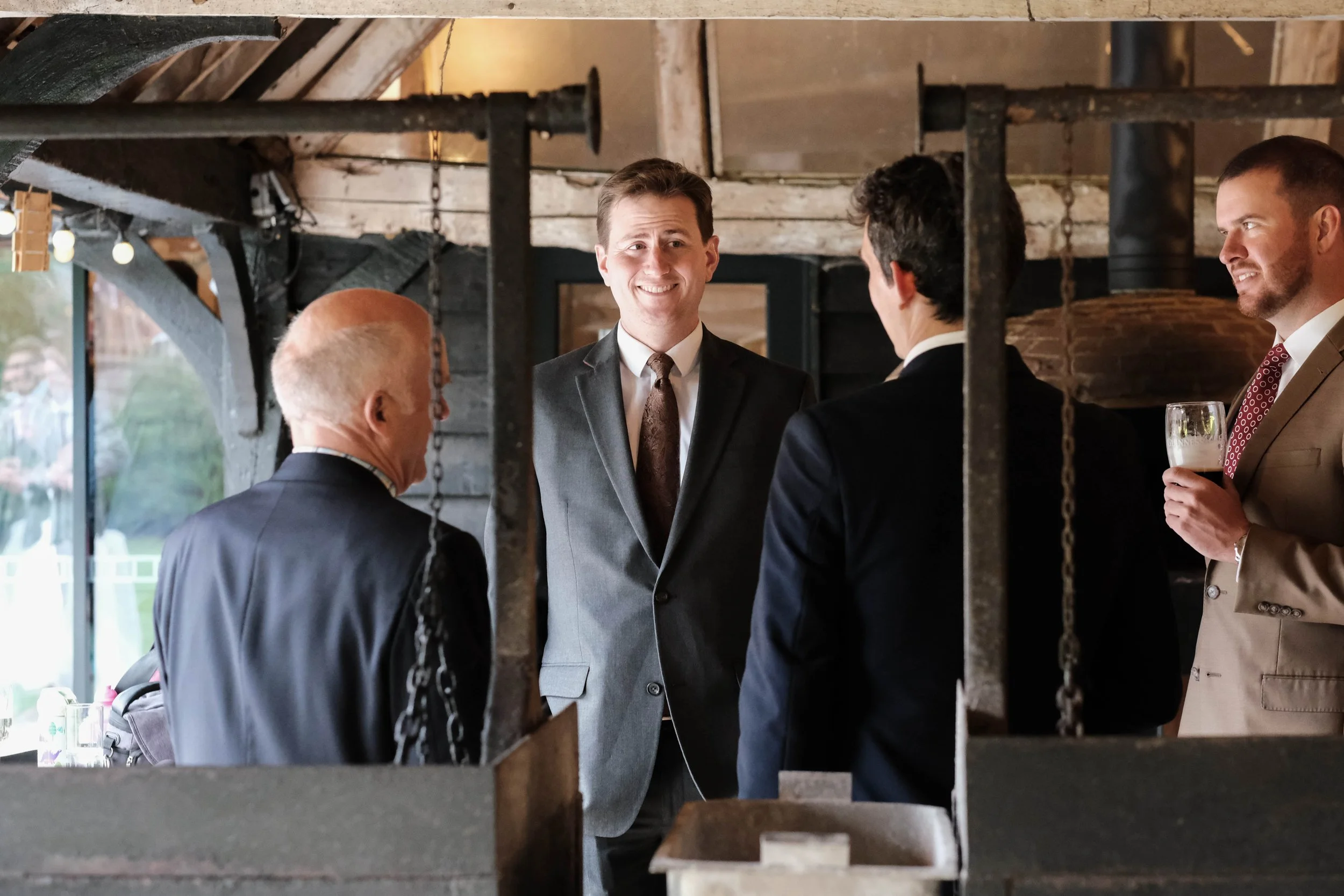 Four men in suits engaging in conversation at a social gathering in an indoor rustic setting, one holding a drink.
