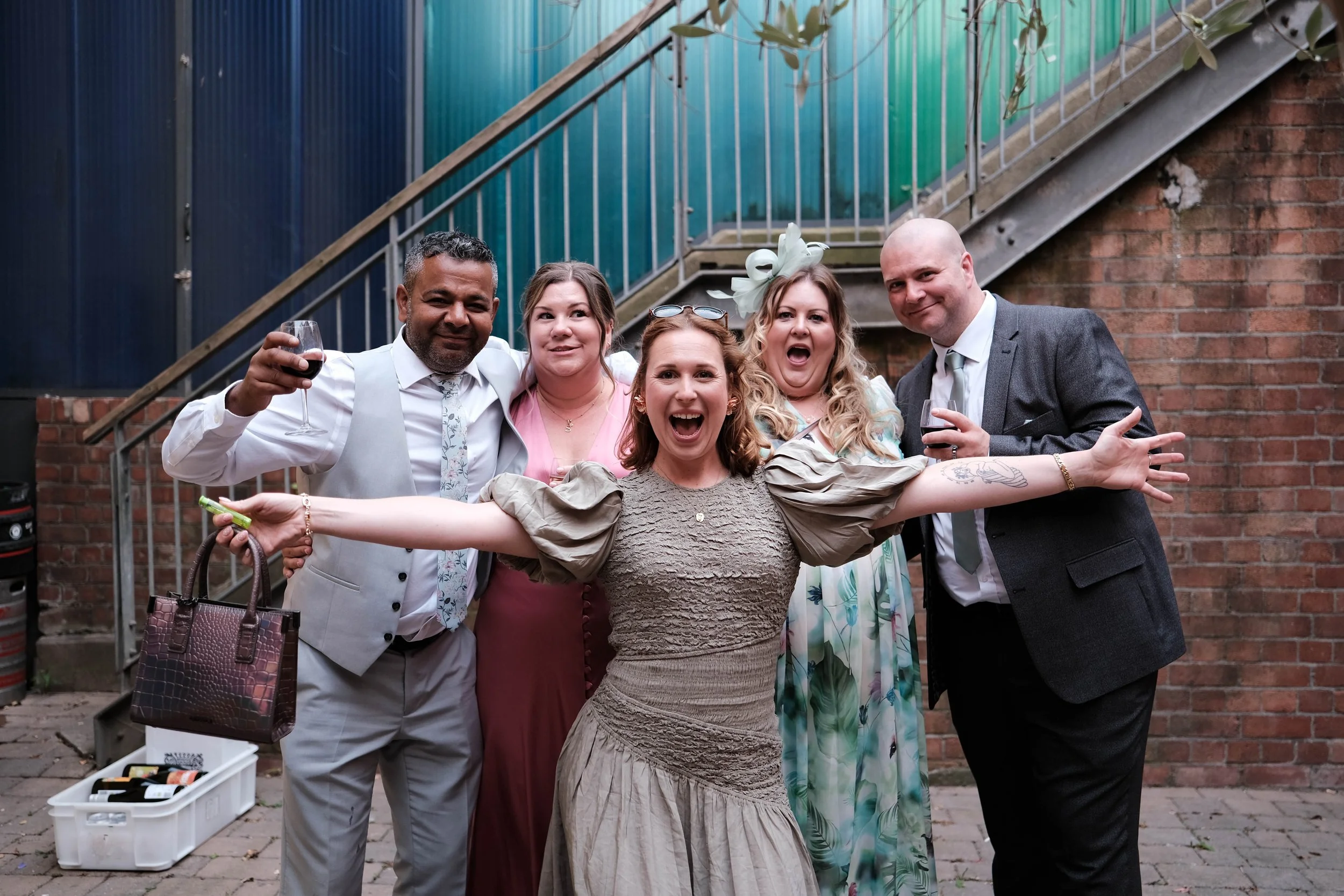 Group of six people smiling and celebrating with drinks at an outdoor event, standing in front of a brick wall and staircase.