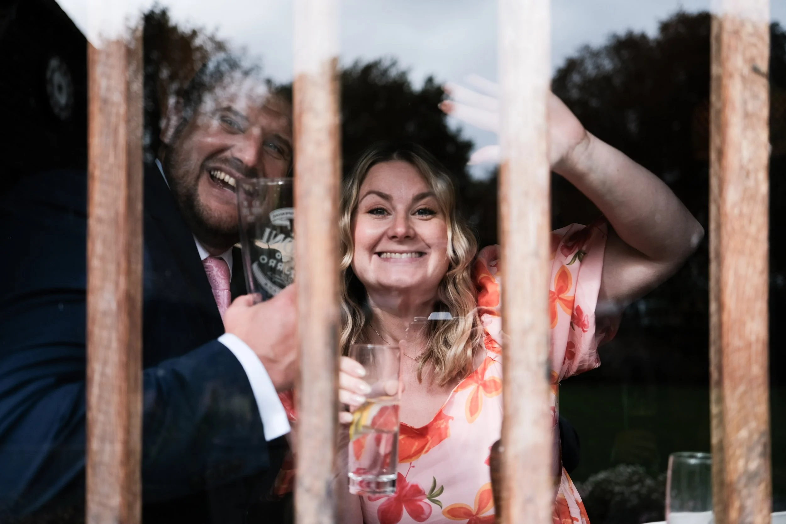 Two people smiling and looking through a window held closed by wooden bars, with one holding a glass of drink.