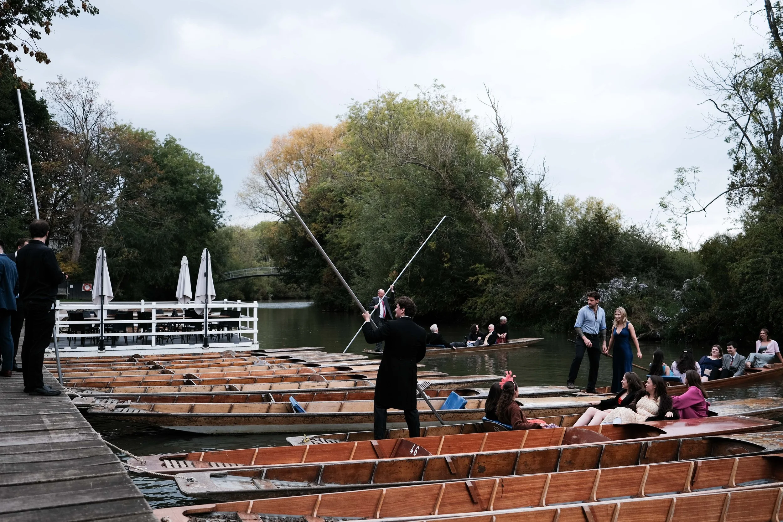 People enjoying a boat ride on a river, with some passengers sitting in boats and others preparing to board, surrounded by trees with autumn foliage.