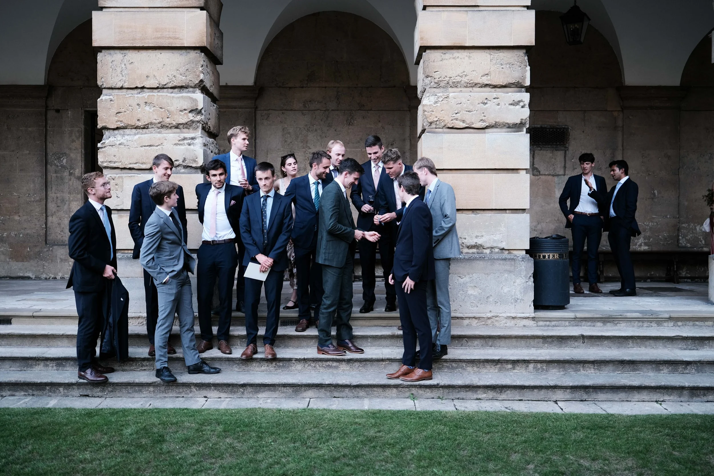 Group of men and women in formal business attire gathered on stairs outside a stone building, some engaging in conversation and looking at a mobile device.