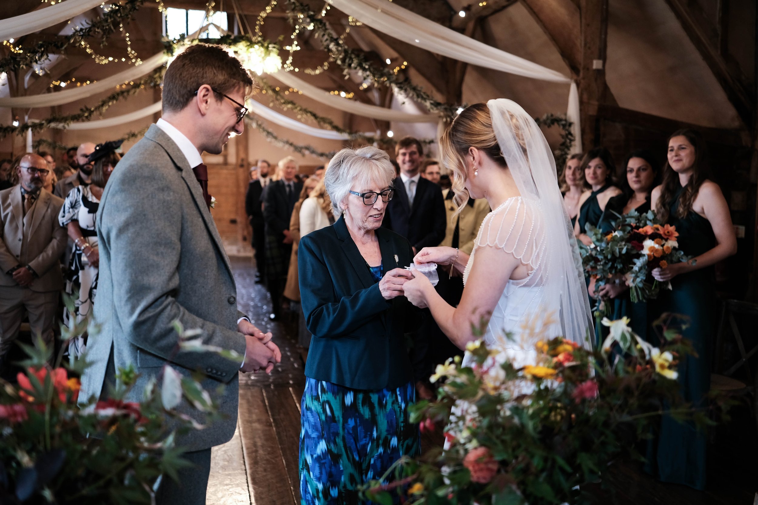 A bride and groom exchanging rings during a wedding ceremony in a rustic decorated venue, with guests in the background.