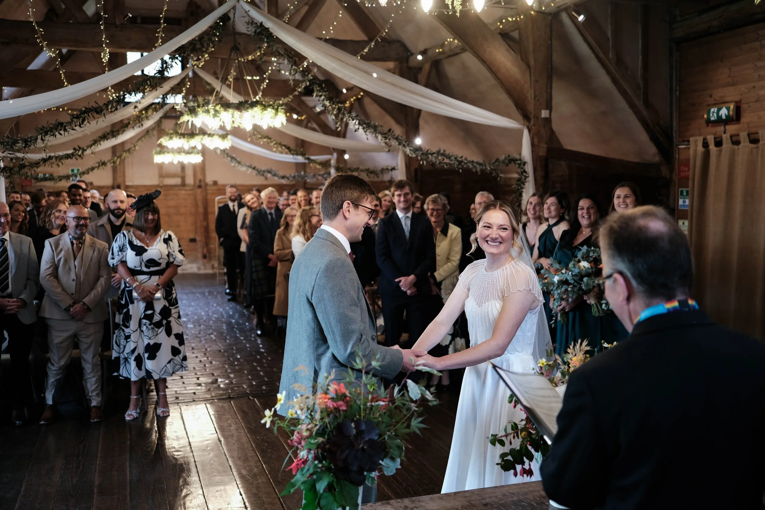 A bride and groom holding hands and smiling during their wedding ceremony in a warmly decorated, rustic wooden hall with guests watching and celebrating.
