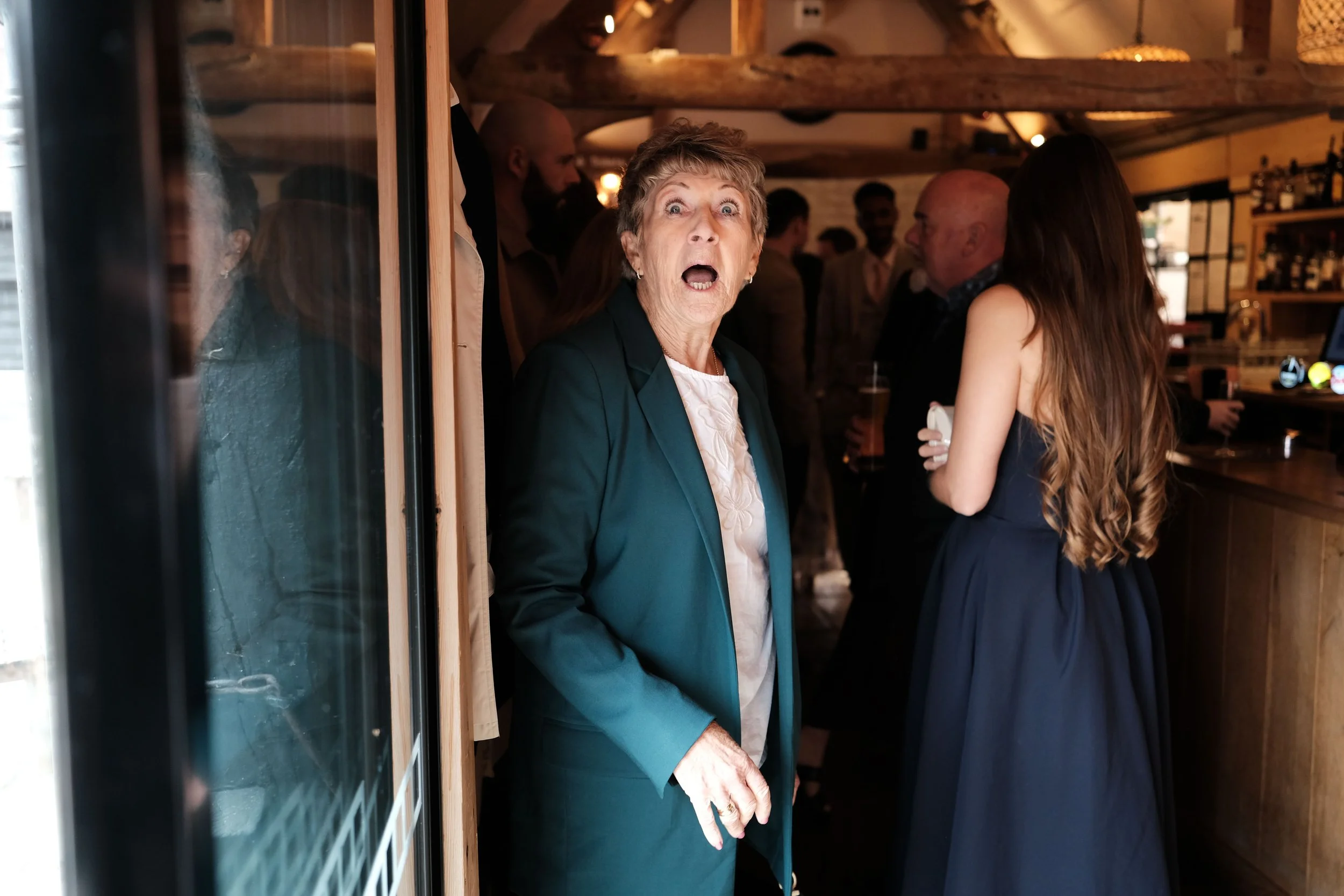 An elderly woman with short, curly hair and a teal blazer looks surprised or shocked at a social gathering inside a wooden-paneled room, with several people in the background.