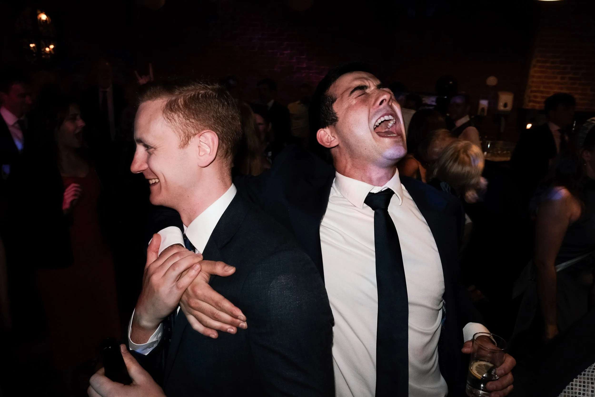 Two men in suits and ties are enjoying a moment of celebration or joy at a festive event, with one holding a drink and both laughing.