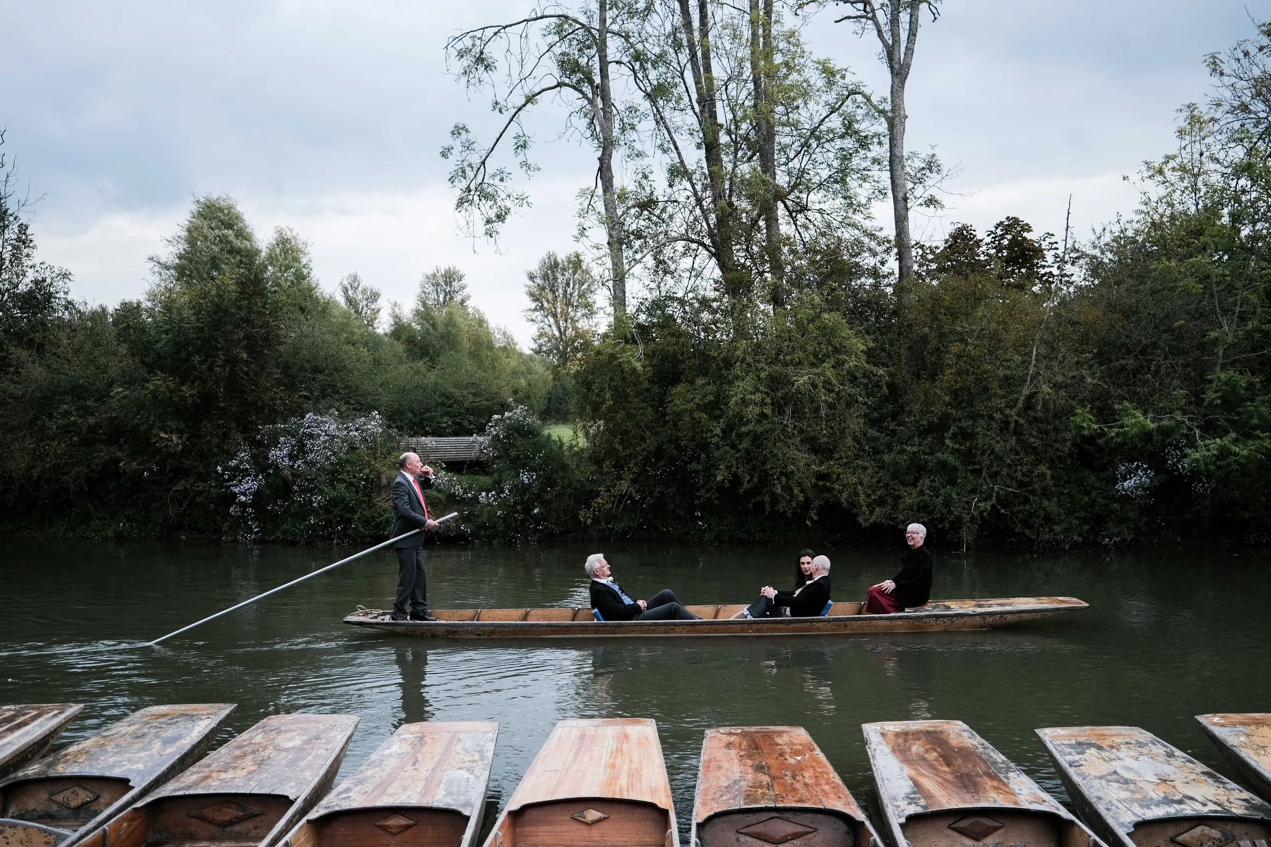 A boat on a river with five people, three seated and two standing, surrounded by greenery and trees, with the boat dock visible in the foreground.