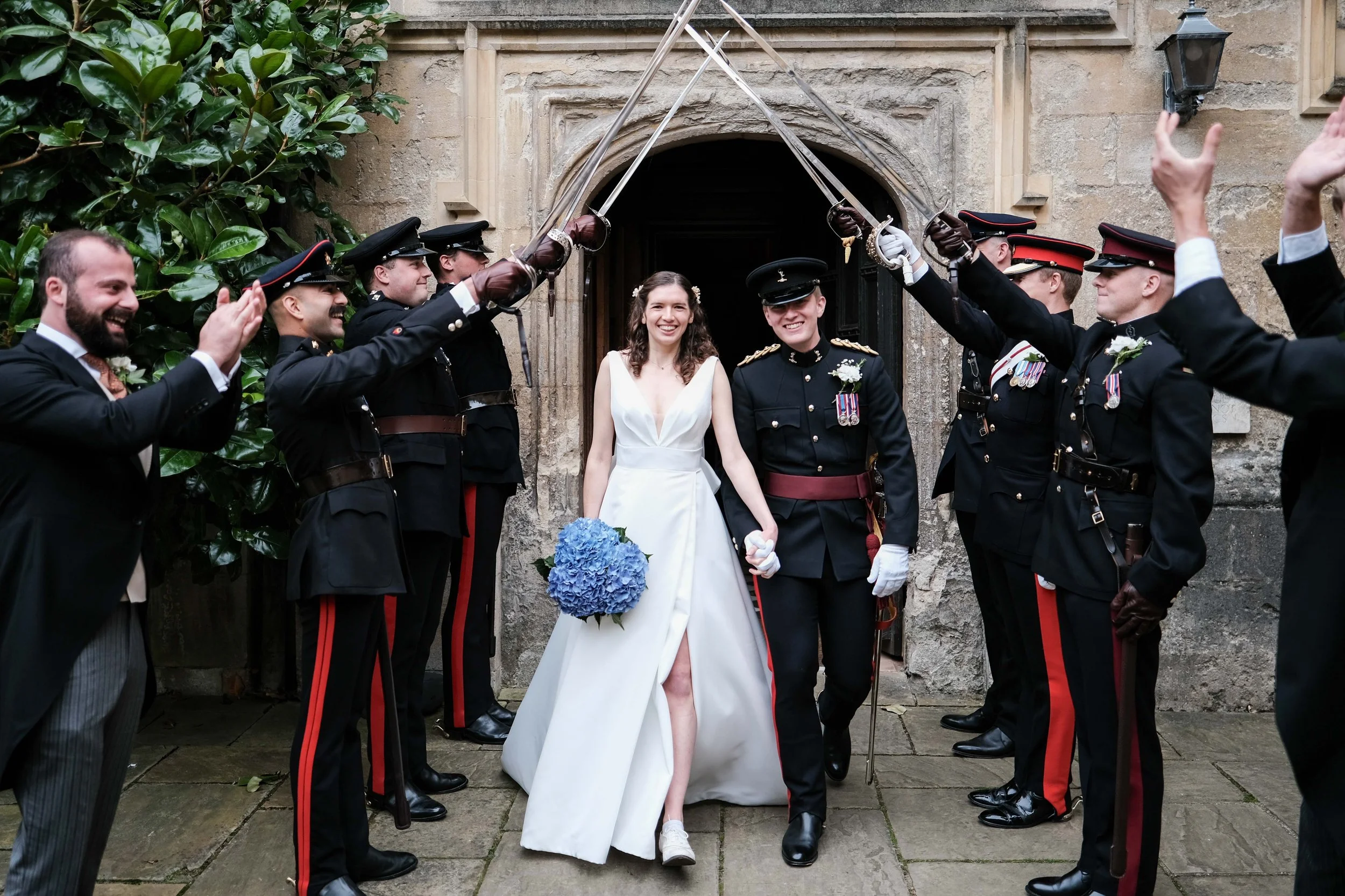 A bride and groom walking through a paper sword arch during their wedding celebration, surrounded by military personnel in uniform.