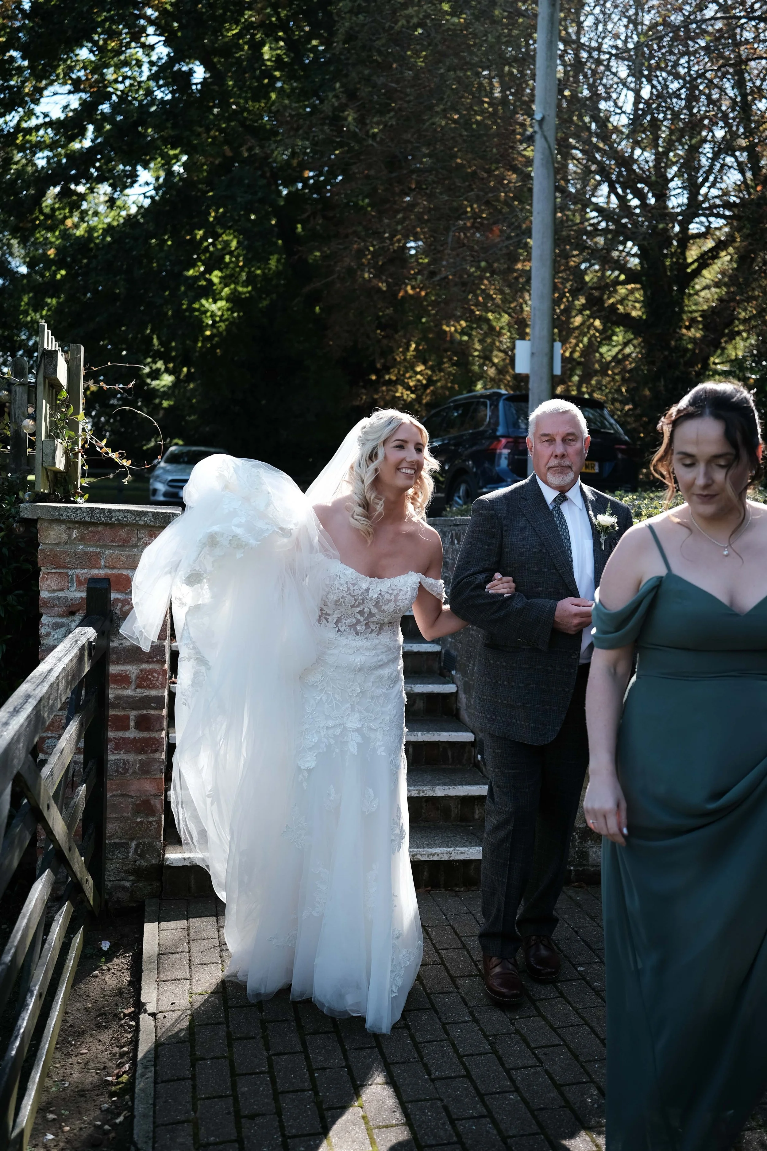 A bride in a wedding dress smiling as she walks outdoors, assisted by an older man, with a woman in a green dress walking beside them. There are trees and parked cars in the background.