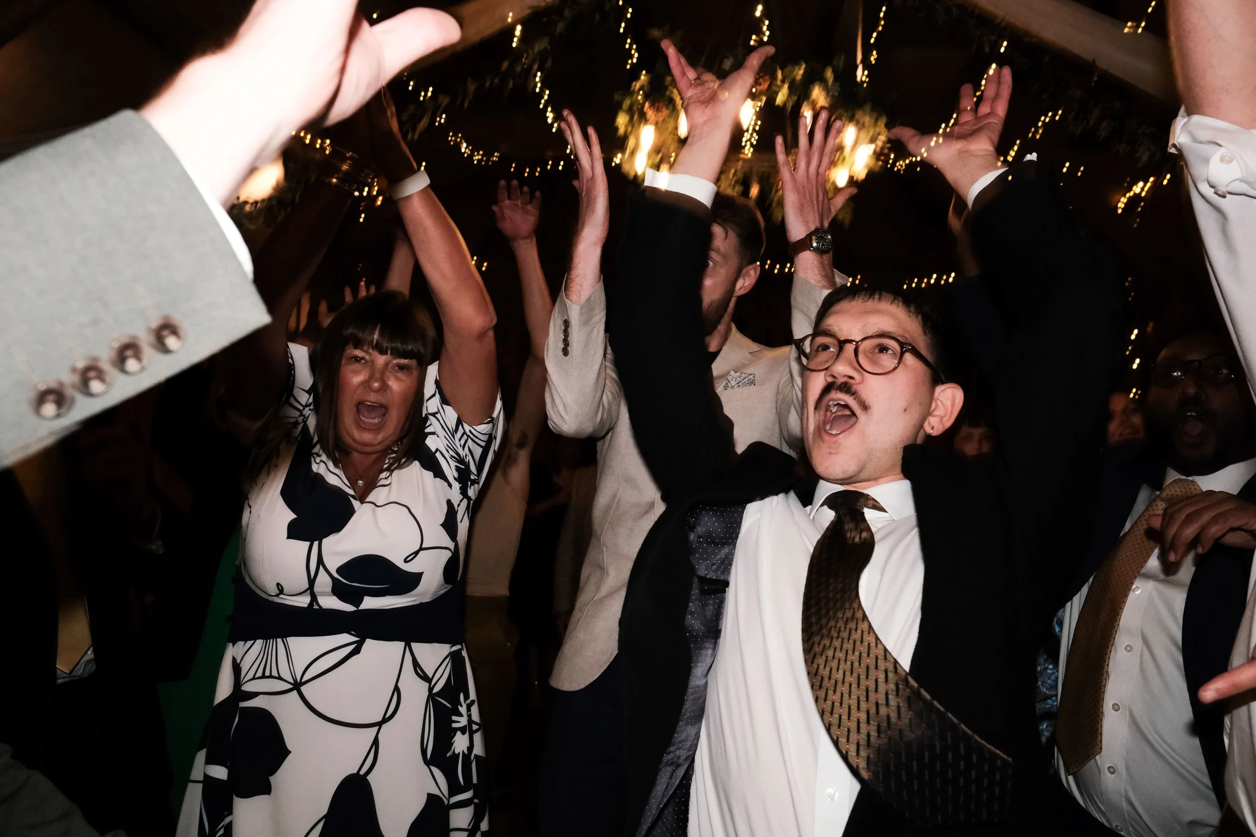 People celebrating at a party with their hands raised, under string lights, in formal attire.