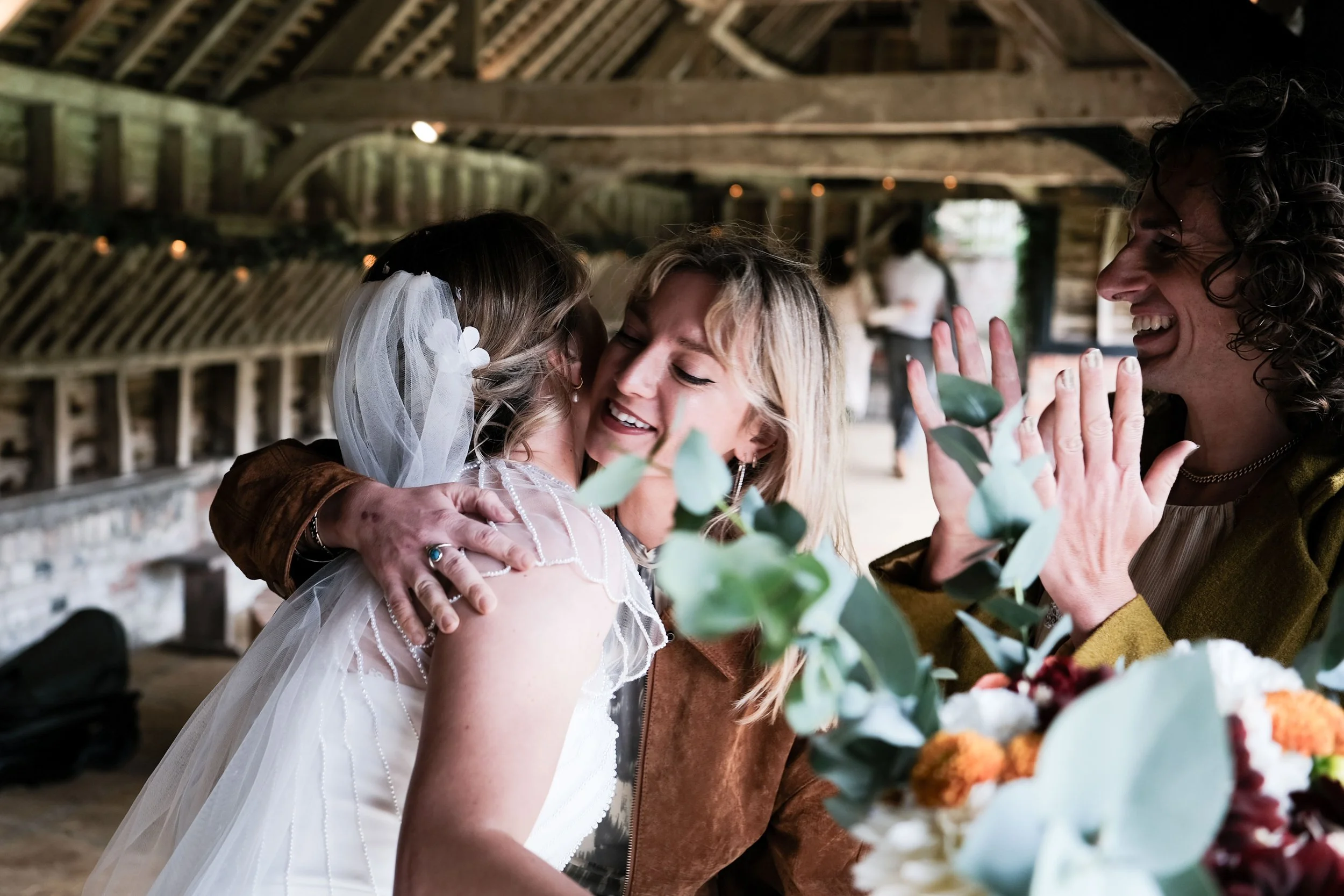 Three women sharing a joyful moment at a wedding, one in a wedding dress, another in a brown jacket, and the third in a mustard yellow blazer, with flowers and greenery in the foreground, in a rustic barn setting.