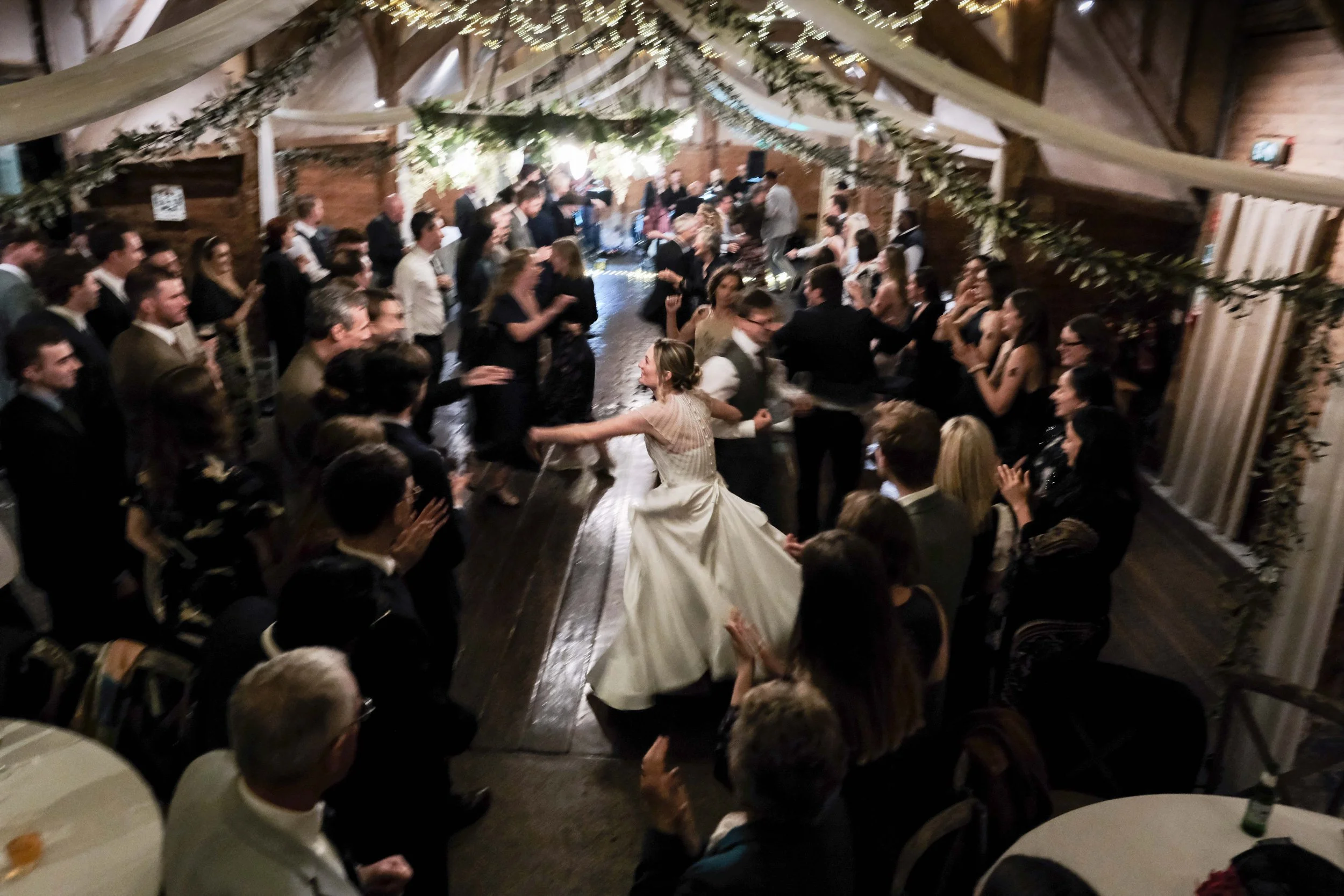 Wedding reception with people dancing in a rustic barn decorated with greenery and string lights.