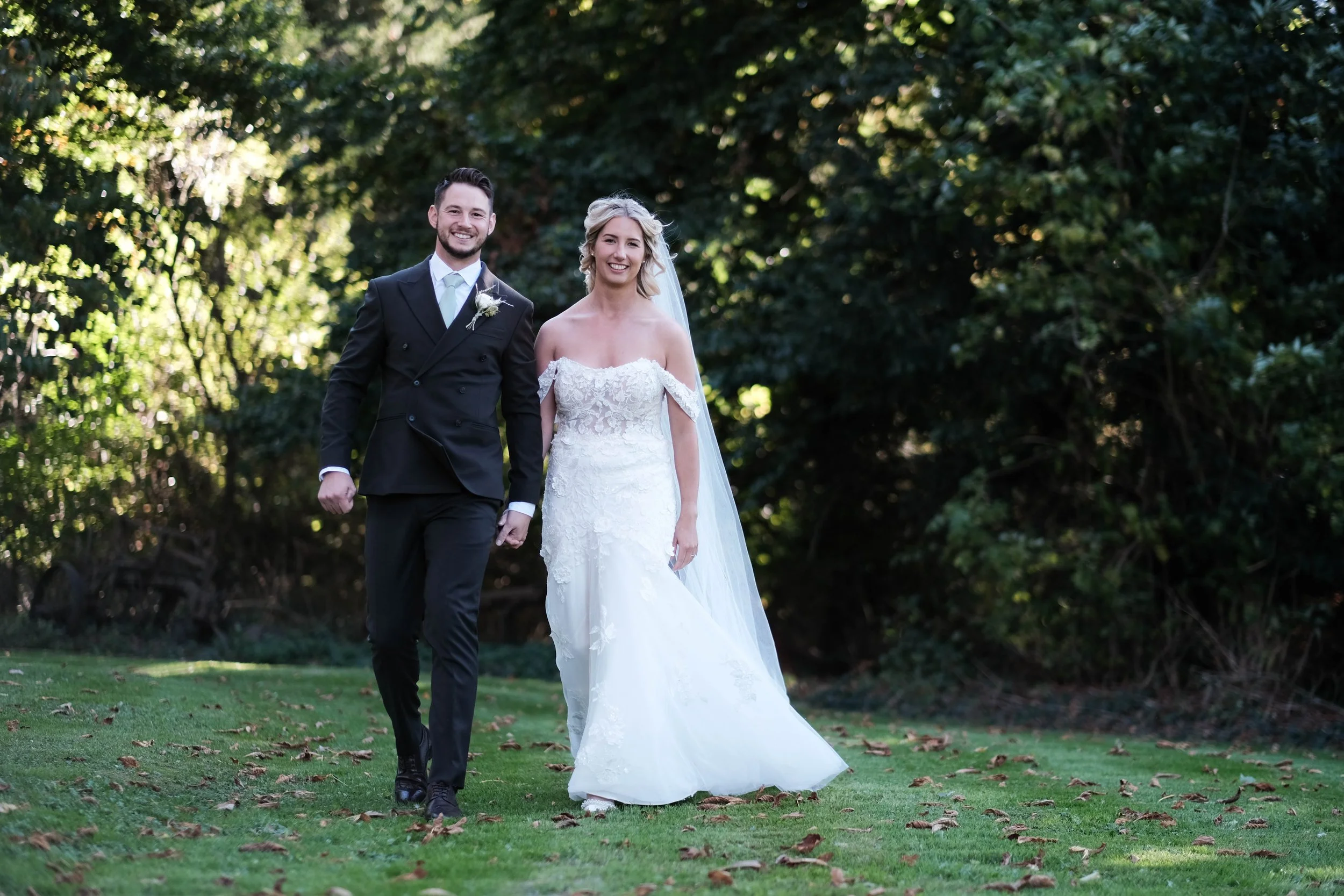 A happy couple walking outdoors on their wedding day, with the bride in a white lace wedding gown and the groom in a black suit, surrounded by green trees and grass.
