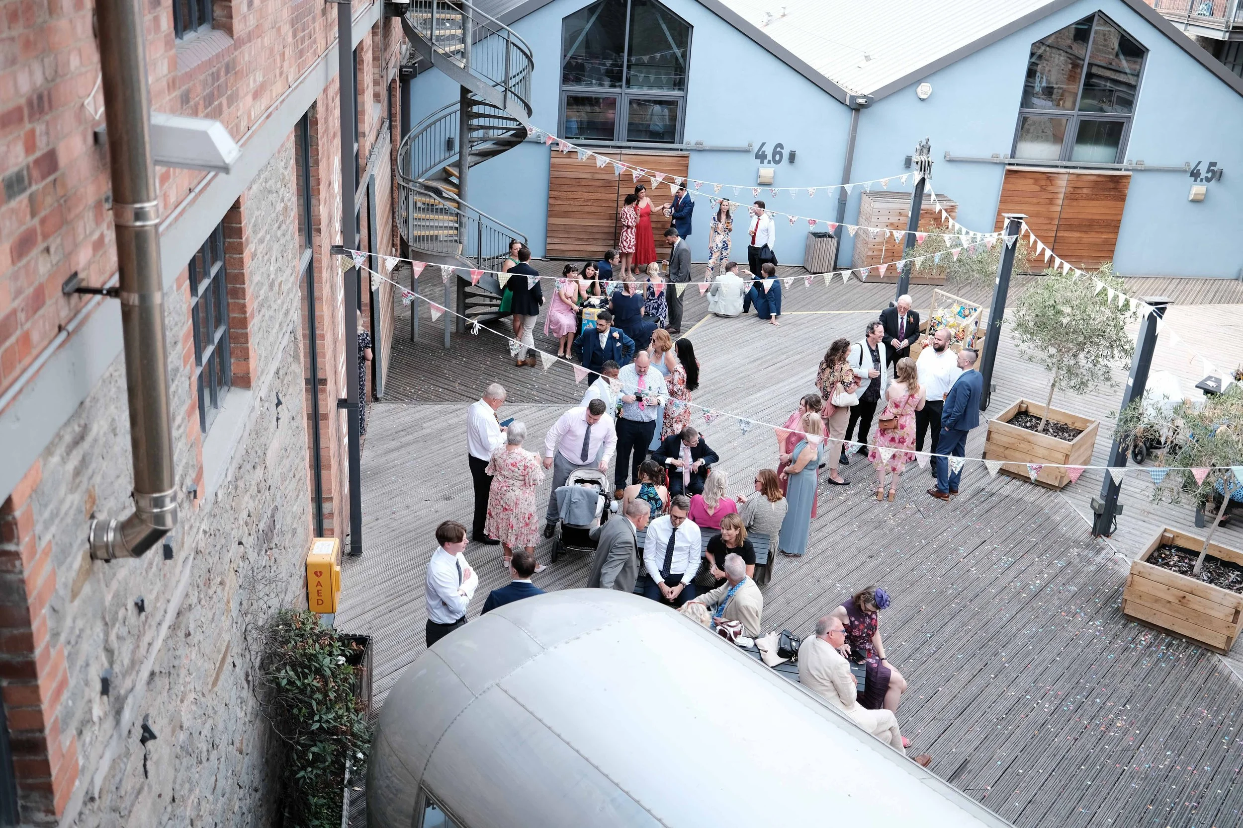 People dressed in formal and semi-formal attire socializing on a decorated outdoor wooden terrace with buntings, potted plants, and a staircase, during a celebration or event.