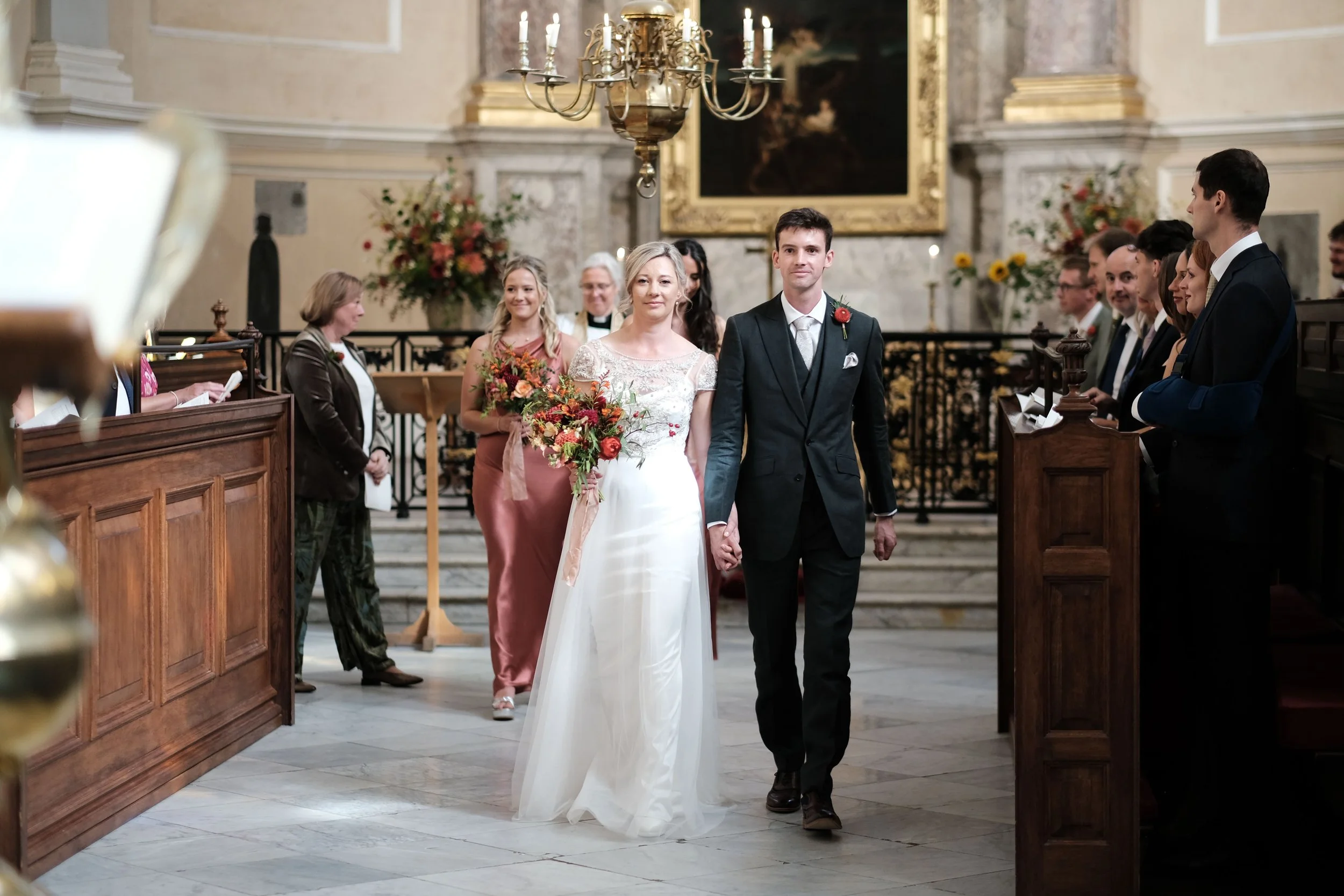 A bride and groom holding hands walking down the aisle of a church during their wedding ceremony, surrounded by guests and floral arrangements.