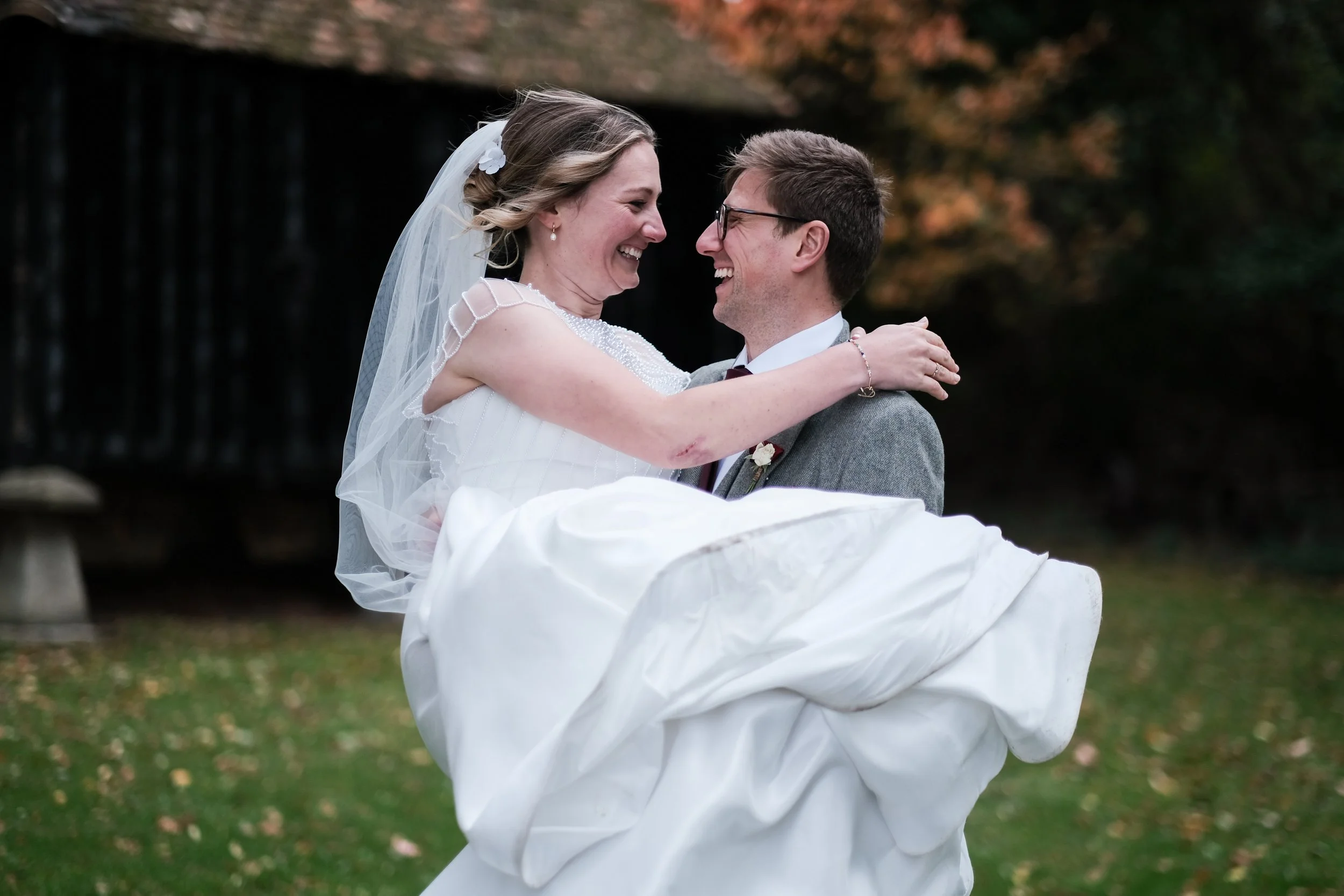 A bride in a white wedding dress and veil is lifted into the air by a groom in a gray suit and glasses, both smiling and looking at each other outdoors.