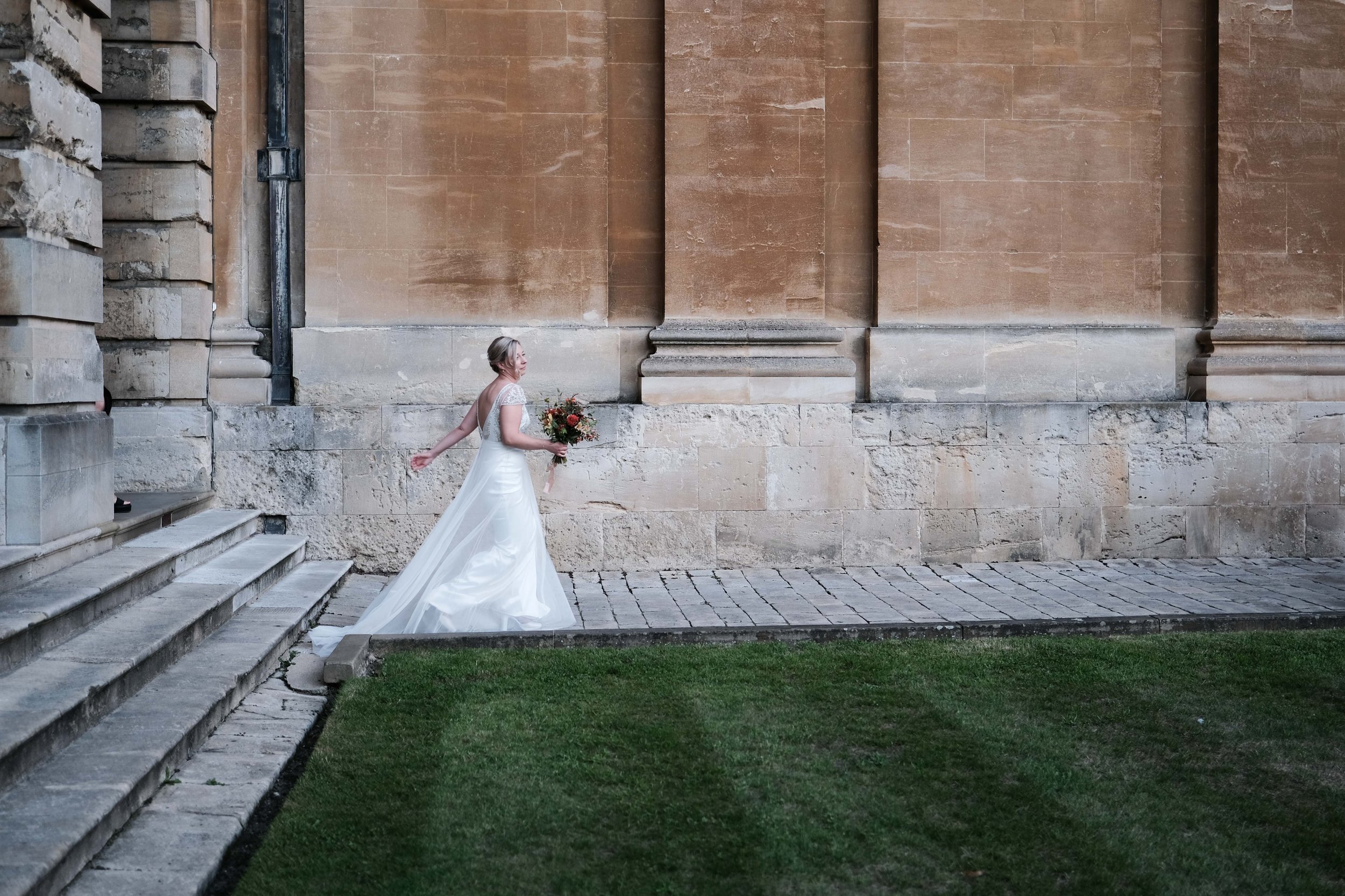 A bride in a white wedding gown holding a bouquet of flowers walking outside near a stone building with steps and a grassy area.
