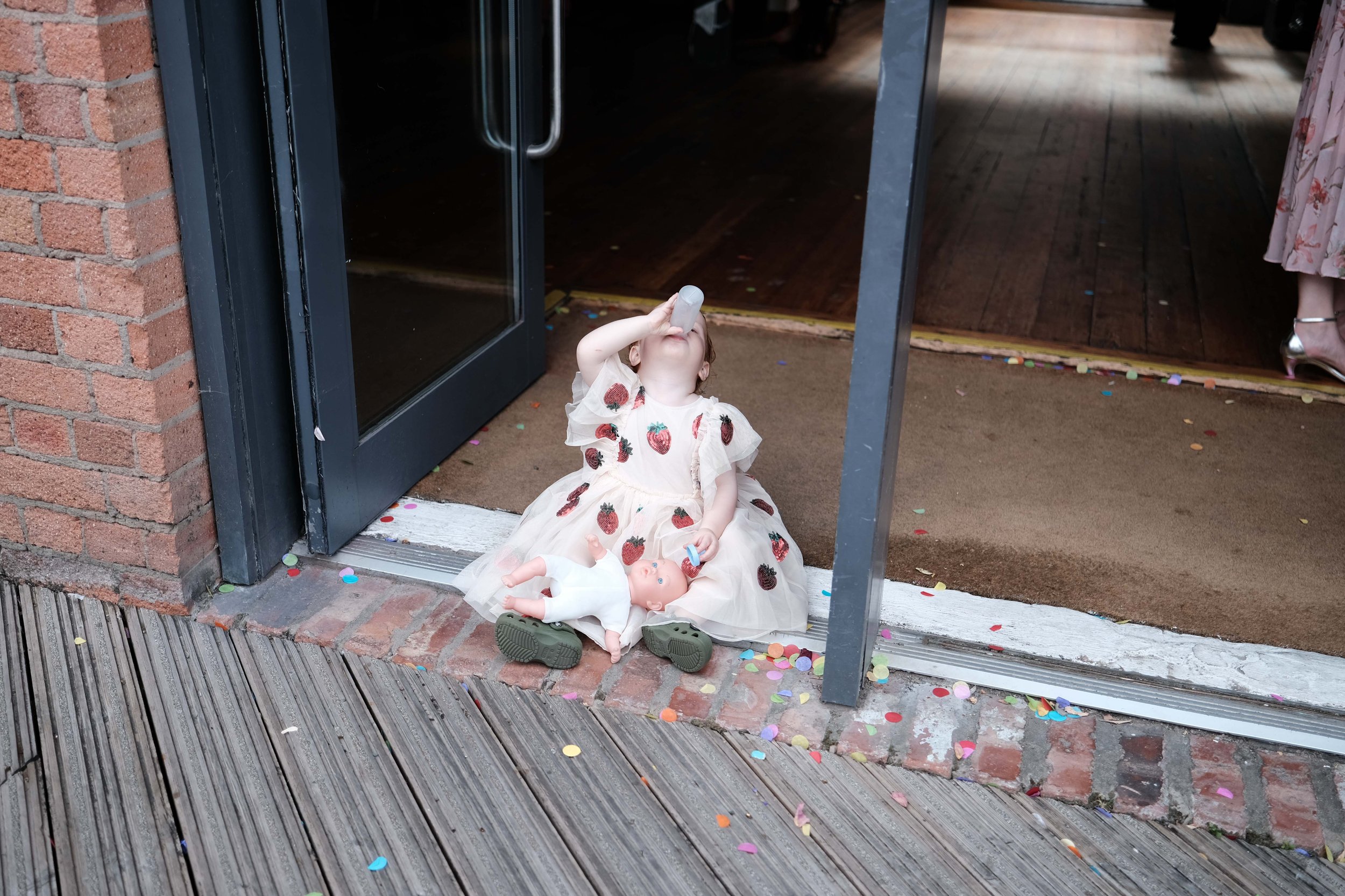 A young girl sitting on the ground outside a building, wearing a dress with strawberry prints, drinking from a bottle. There is a doll lying next to her, and colorful confetti scattered around.