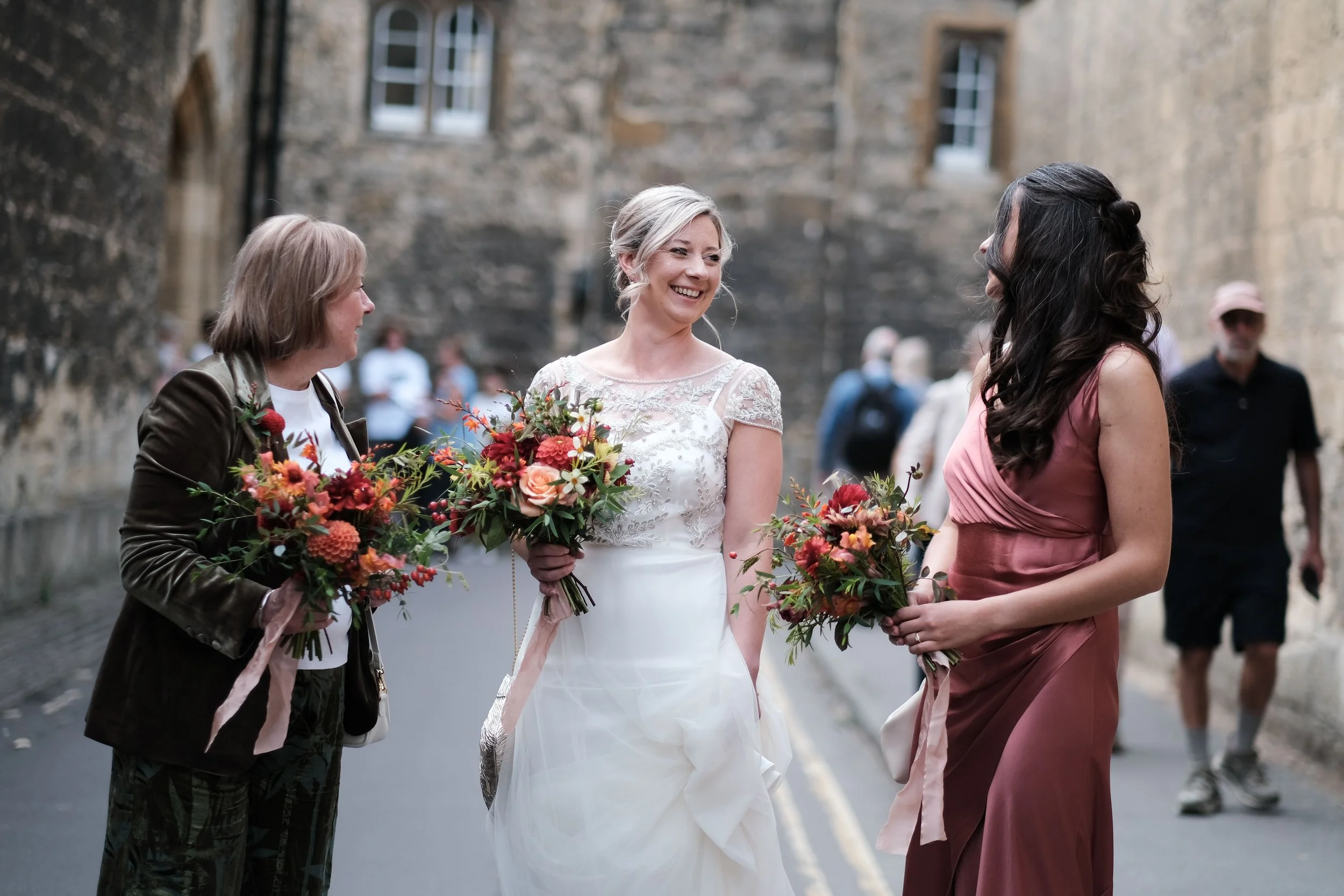 Three women with bouquets of flowers chatting and smiling in a street, with people in the background.