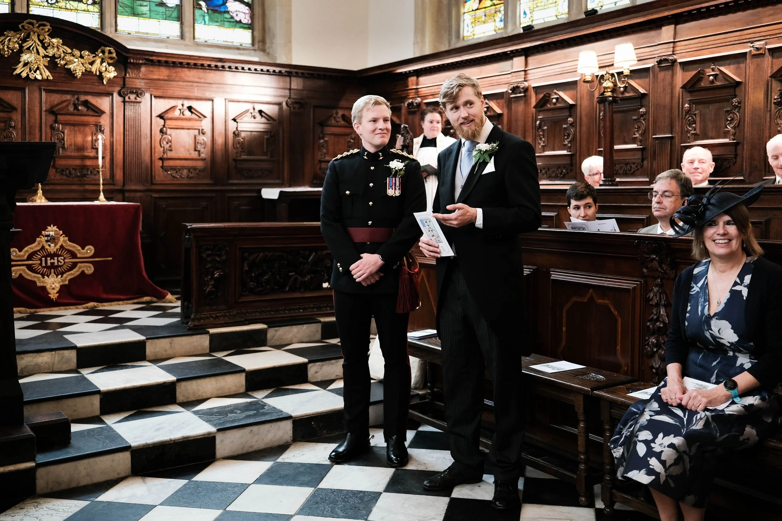 A wedding ceremony inside a church with two men standing together, one in a military uniform and the other in a wedding suit, surrounded by seated guests.