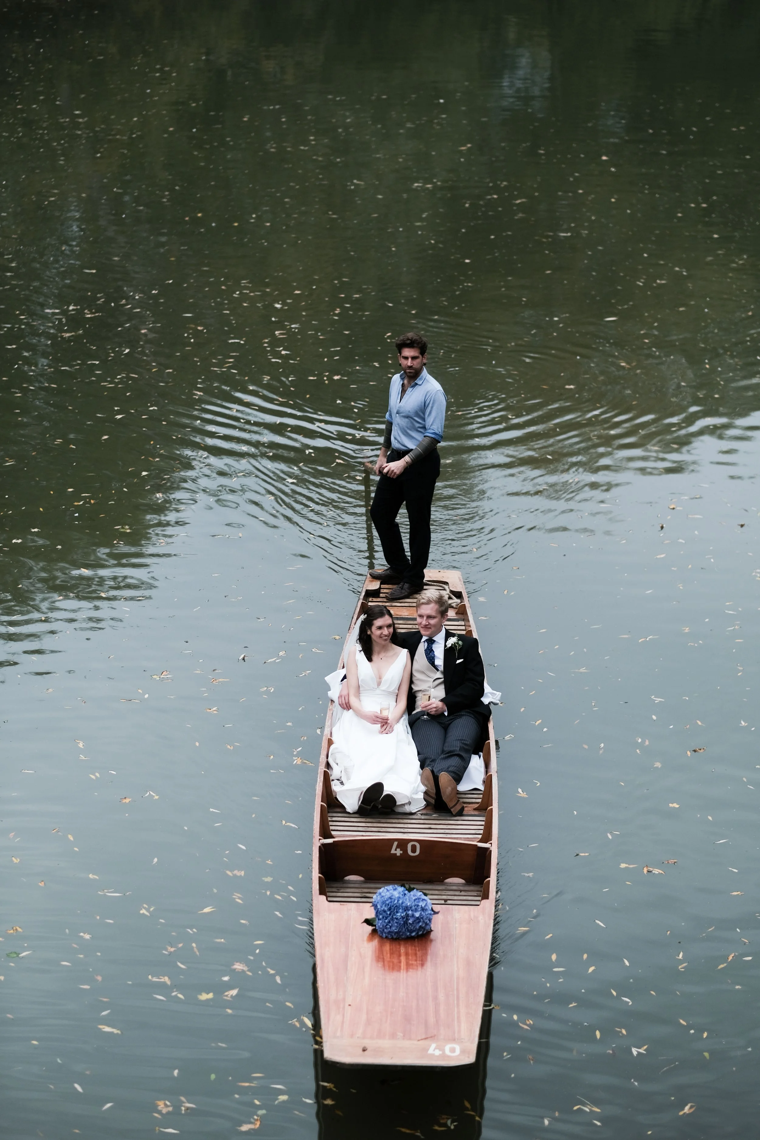 A couple in wedding attire sitting in a boat on a calm lake, with a man standing at the rear of the boat.