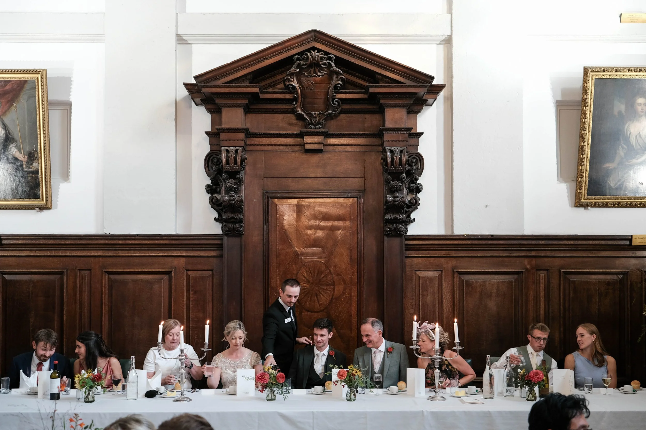 People sitting at a long wedding banquet table with candles, flowers, and drinks, inside a grand room with wood-paneled walls and large paintings.