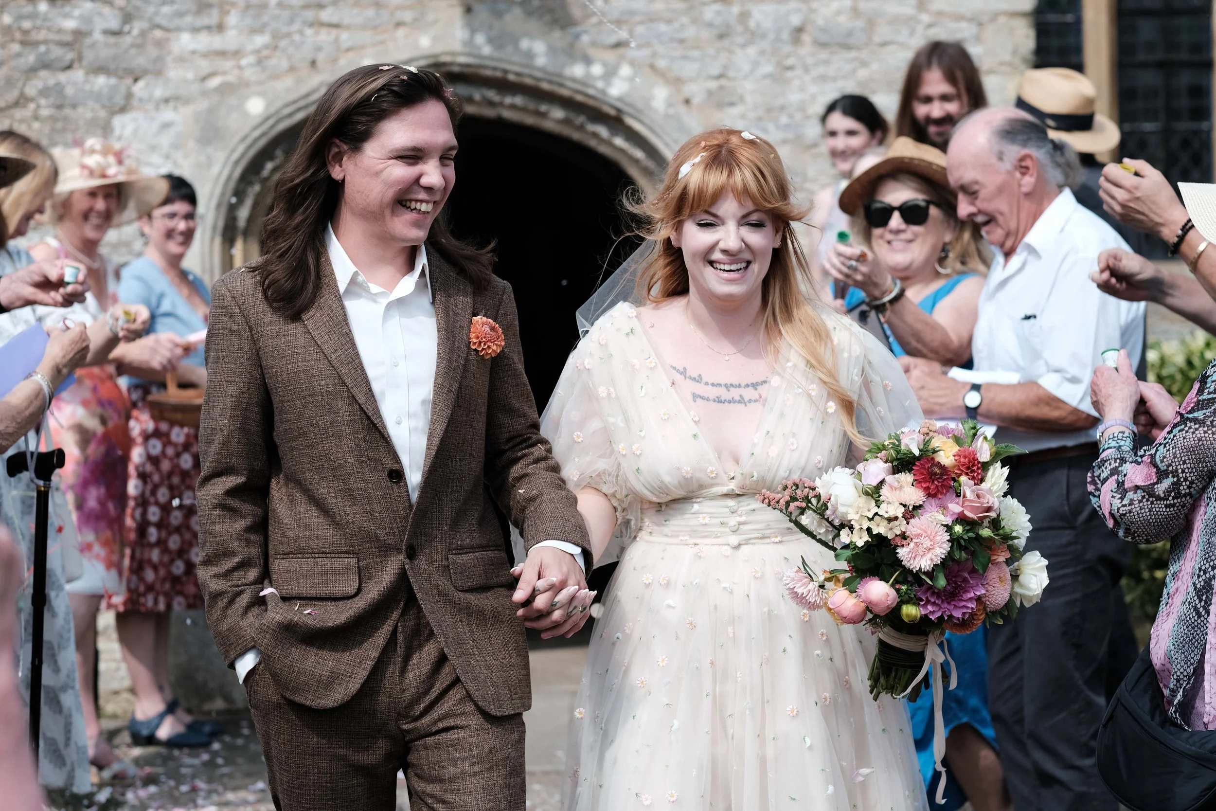 A newlywed couple smiles as they walk hand in hand through a crowd of celebrating guests outside a stone building, with the bride holding a bouquet of flowers.