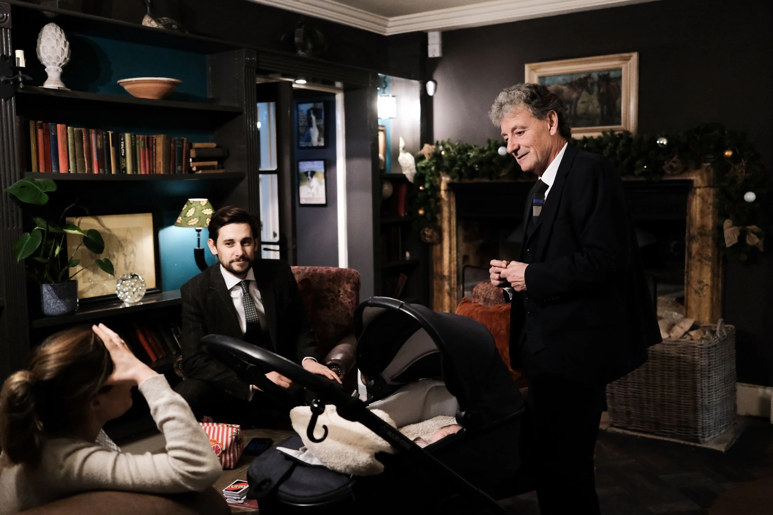 Family gathered in a cozy living room decorated for Christmas, with a fireplace, Christmas garland, and family members sitting and standing around a stroller.