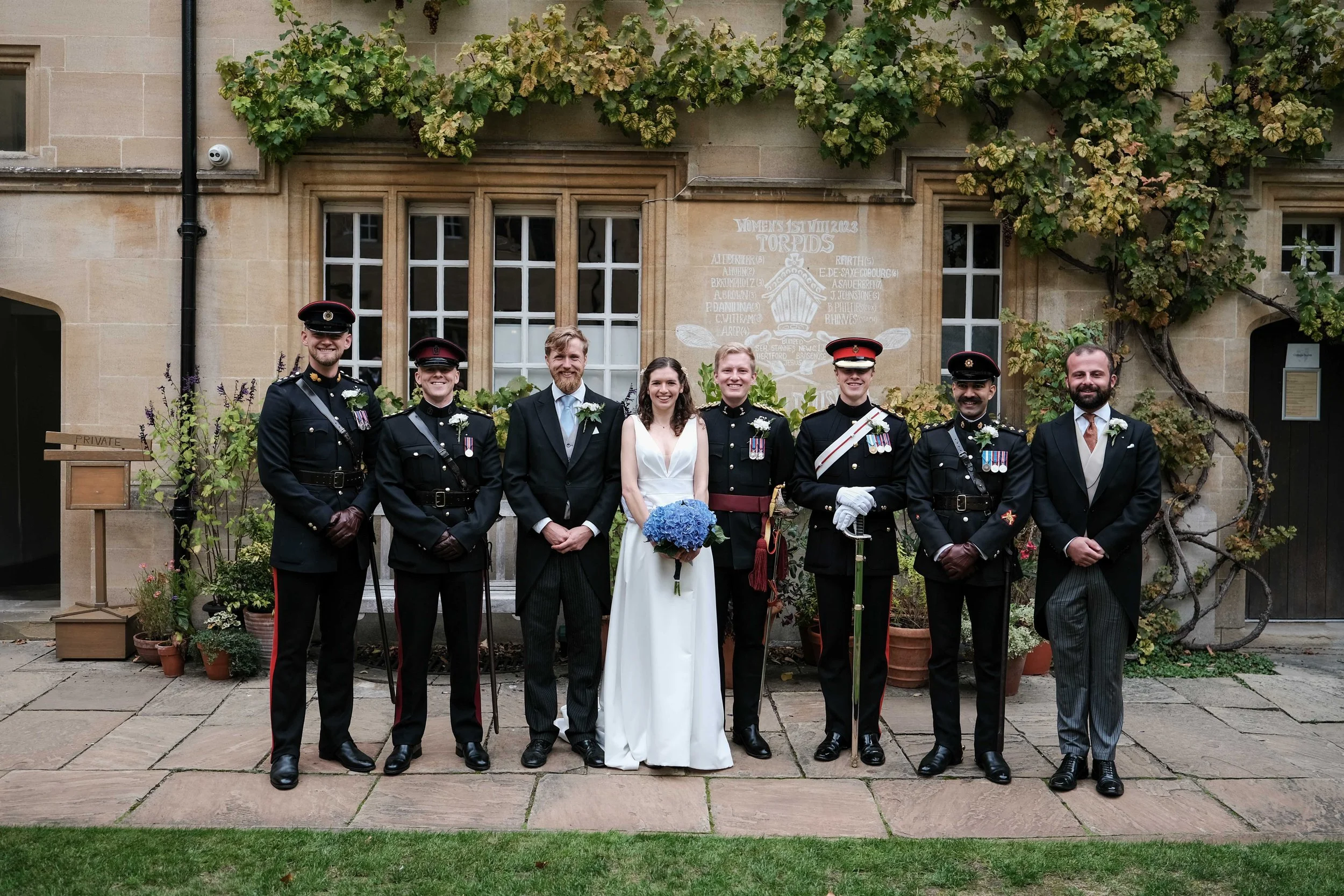 Group of people, including members of the military in uniform and a bride in a white dress holding a bouquet of blue flowers, posing for a photo outside a stone building with greenery.