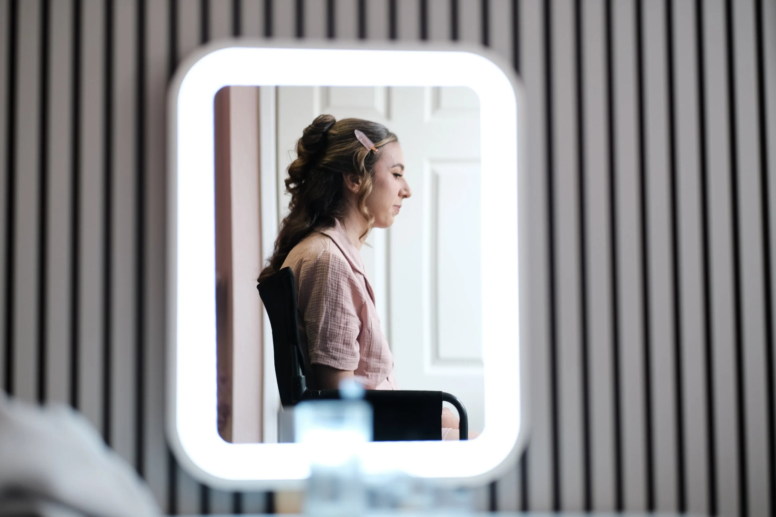Side view of a woman sitting in a chair, looking down, with her hair partially pinned back, reflected in a backlit mirror.
