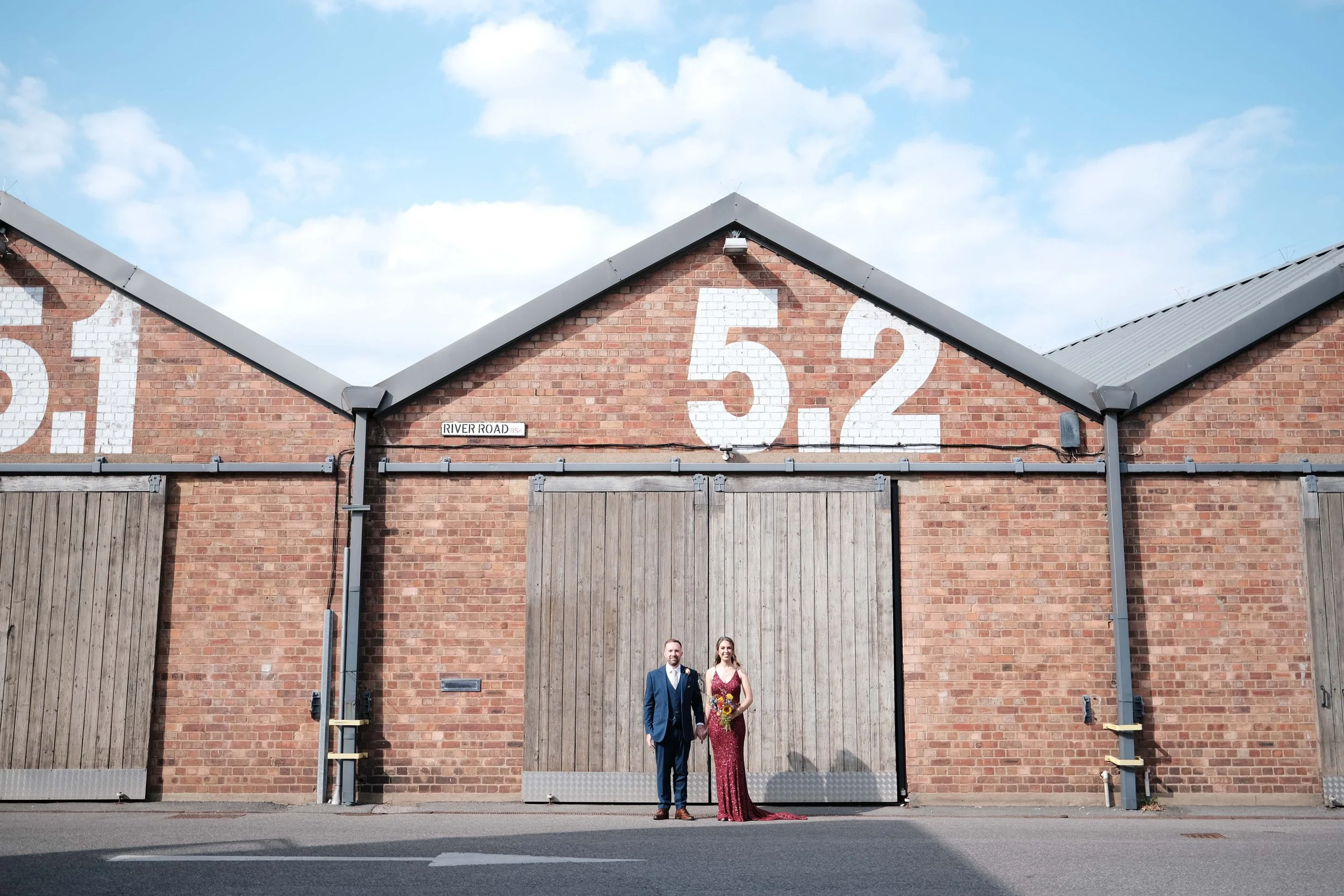 A bride and groom standing hand in hand in front of a brick building with large white painted numbers 5-2 and a street sign that reads 'RIVER ROAD.'