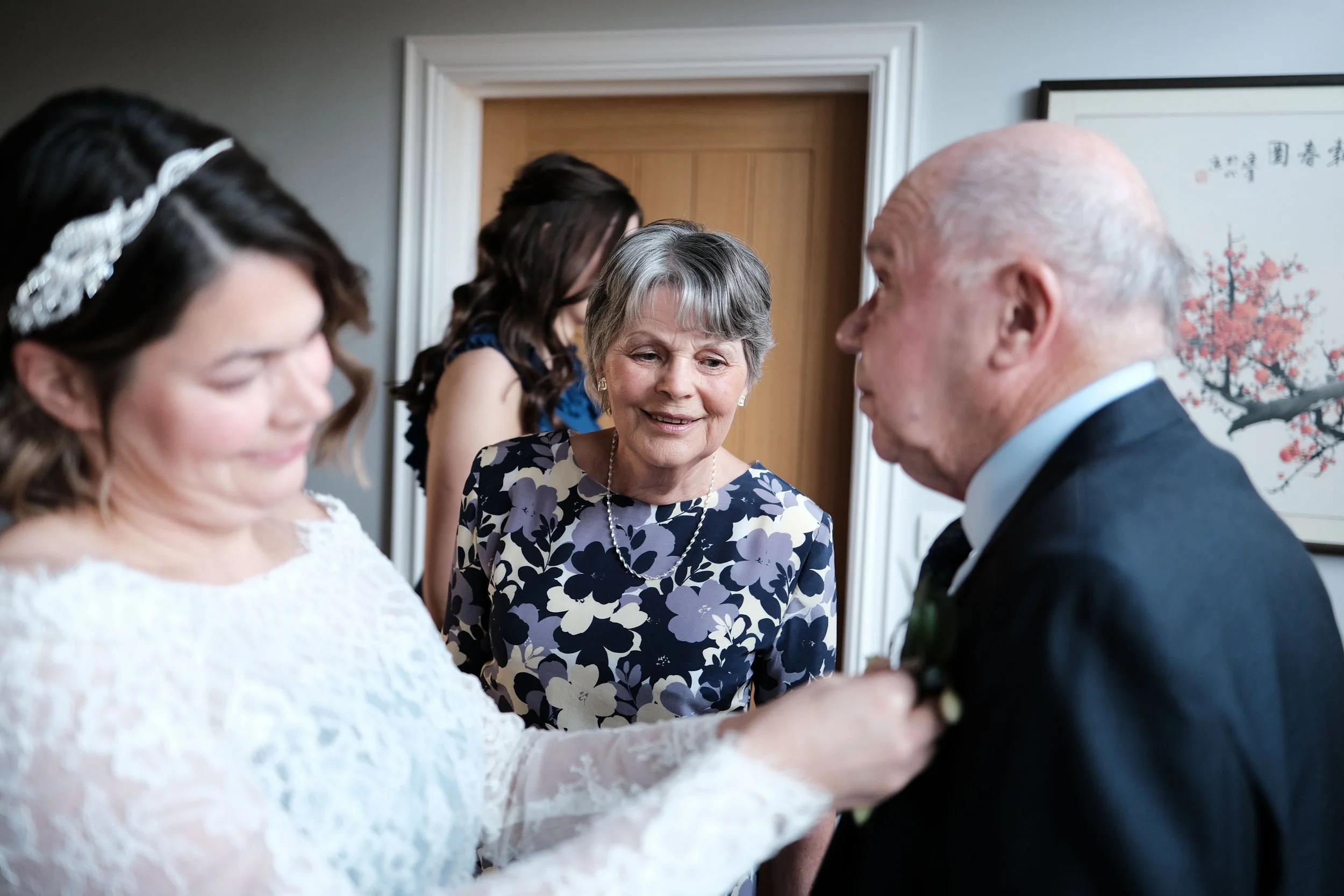 A woman in a white lace dress helps an elderly man in a tuxedo with a boutonniere during a wedding or formal event, with two women in the background and a framed Asian-style themed artwork on the wall.