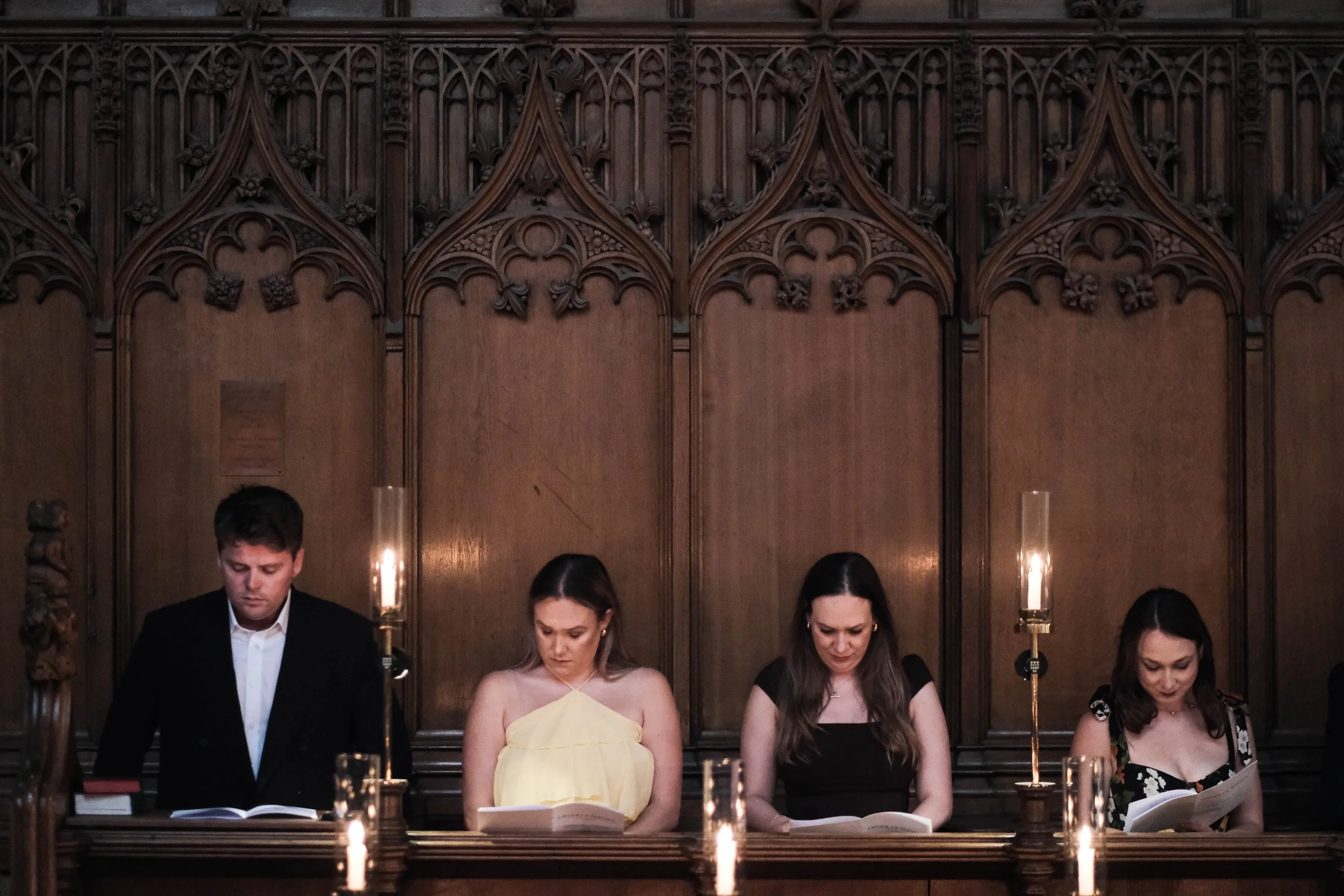 Four people sitting at a wooden bench in a dimly lit room with gothic-style woodwork and candlesticks, reading from books.