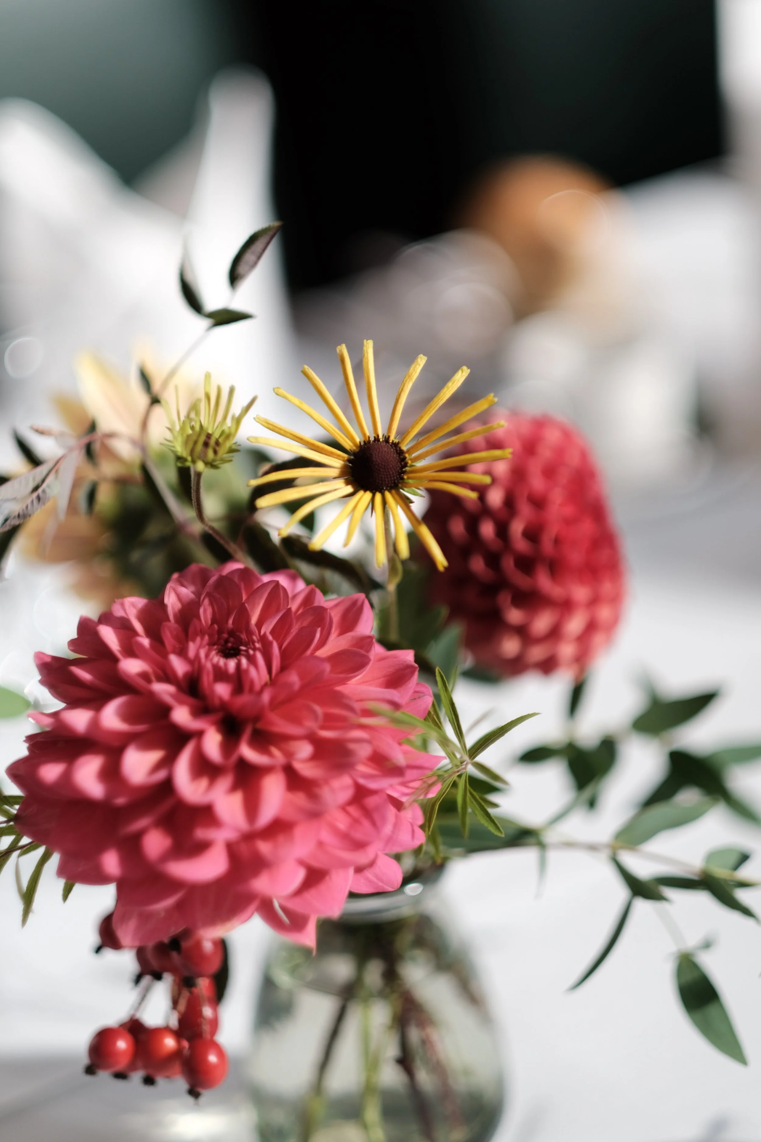 A close-up of a floral arrangement featuring pink, yellow, and reddish flowers in a glass vase with greenery.
