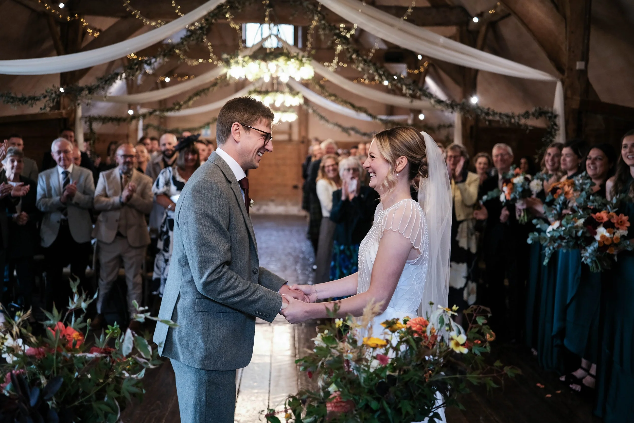 A bride and groom holding hands and smiling at each other during their wedding ceremony inside a decorated barn, with family and friends clapping and watching in the background.