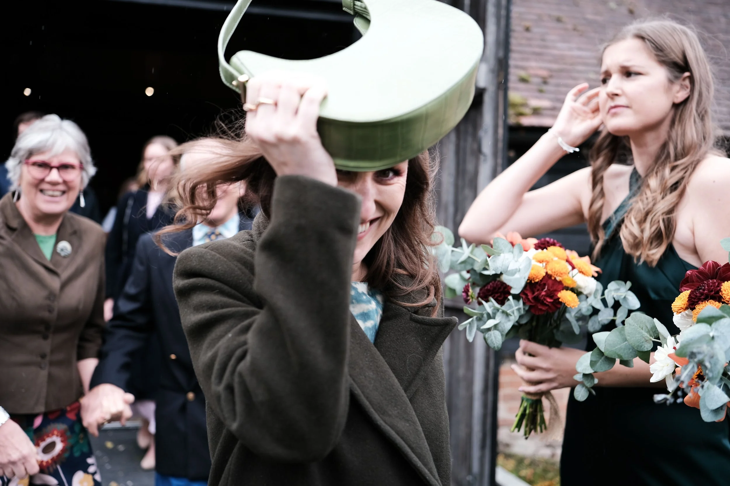 A woman smiling and playfully holding a green watering can over her head while surrounded by a group of women, some holding flowers, outdoors near a rustic building.