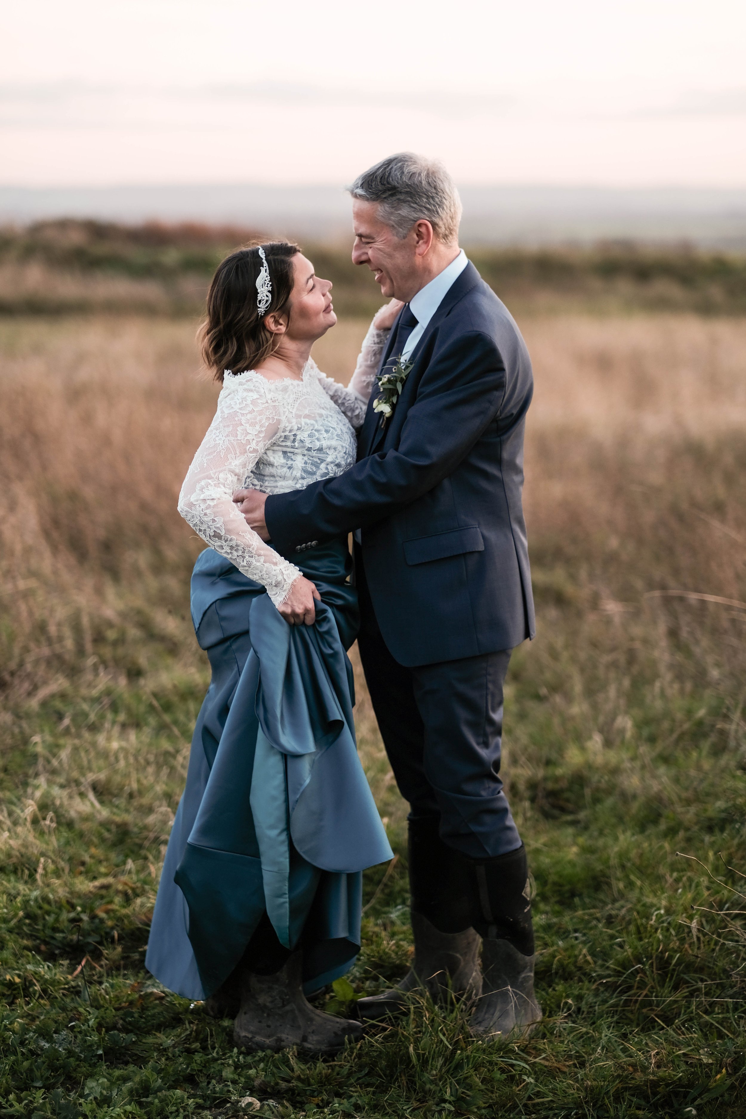A couple in wedding attire standing in a grassy field, smiling and holding each other, with the woman lifting her dress slightly, during sunset.
