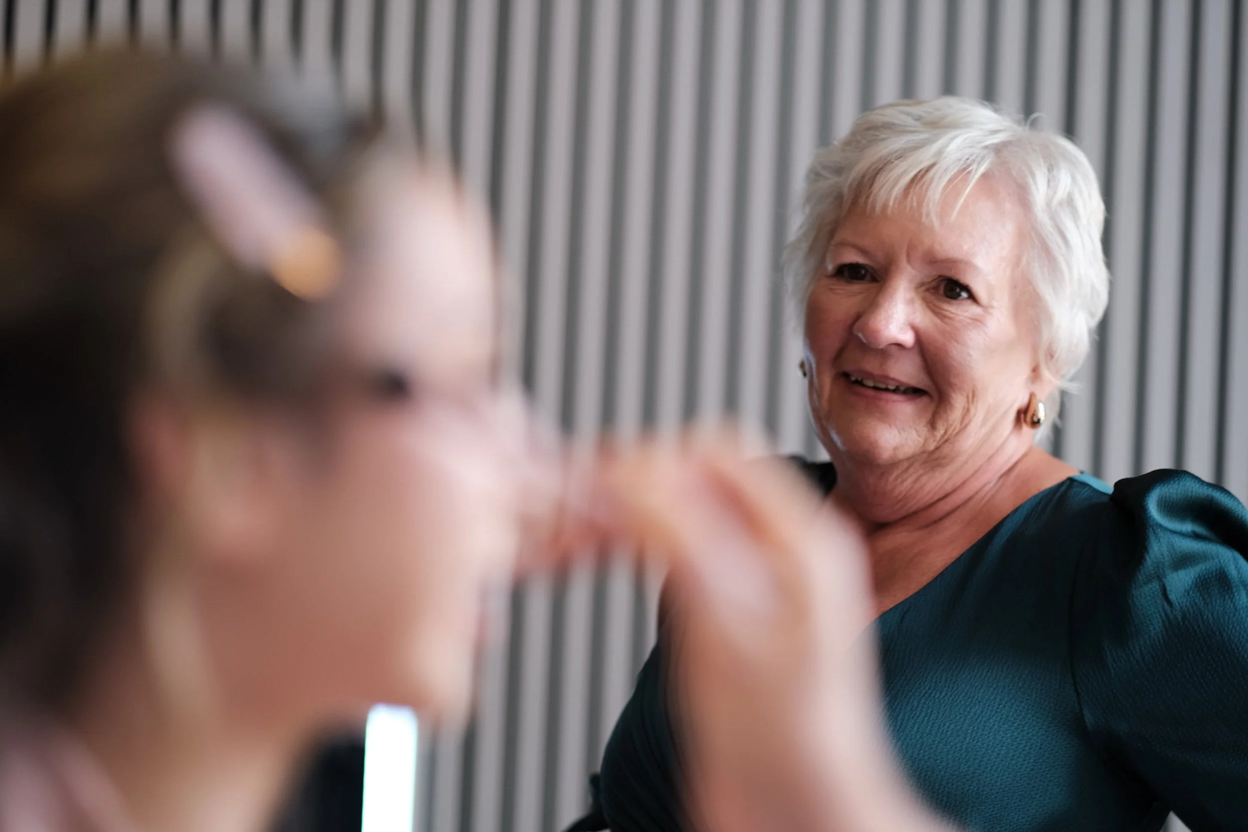 An elderly woman with short white hair smiling and looking at a young woman with glasses, who is slightly out of focus in the foreground.