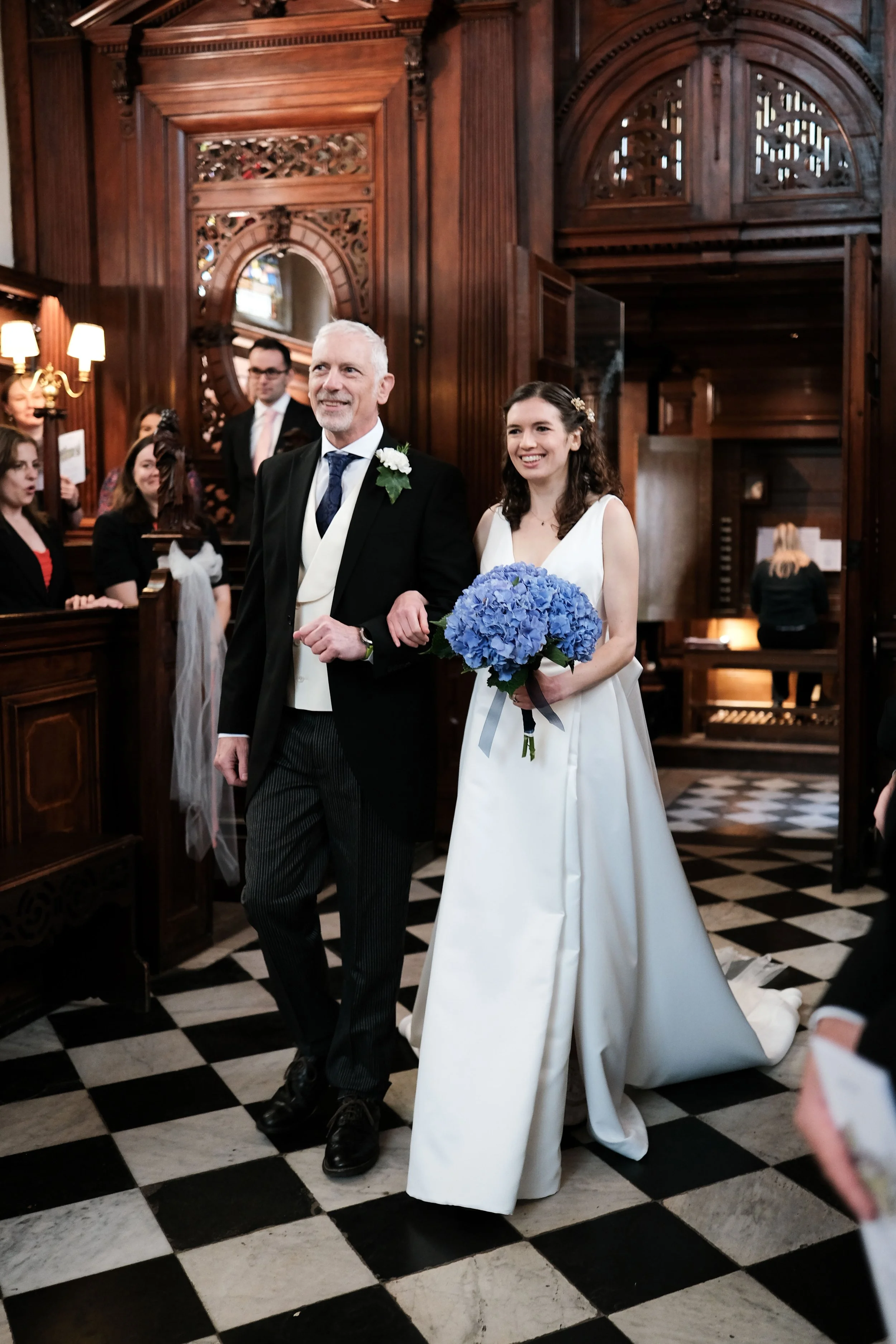 A bride walking down the aisle with her father during a wedding ceremony inside a church. The bride is holding a bouquet of blue flowers and is wearing a white wedding dress. The church has dark wooden paneling and a black and white checkered floor. 