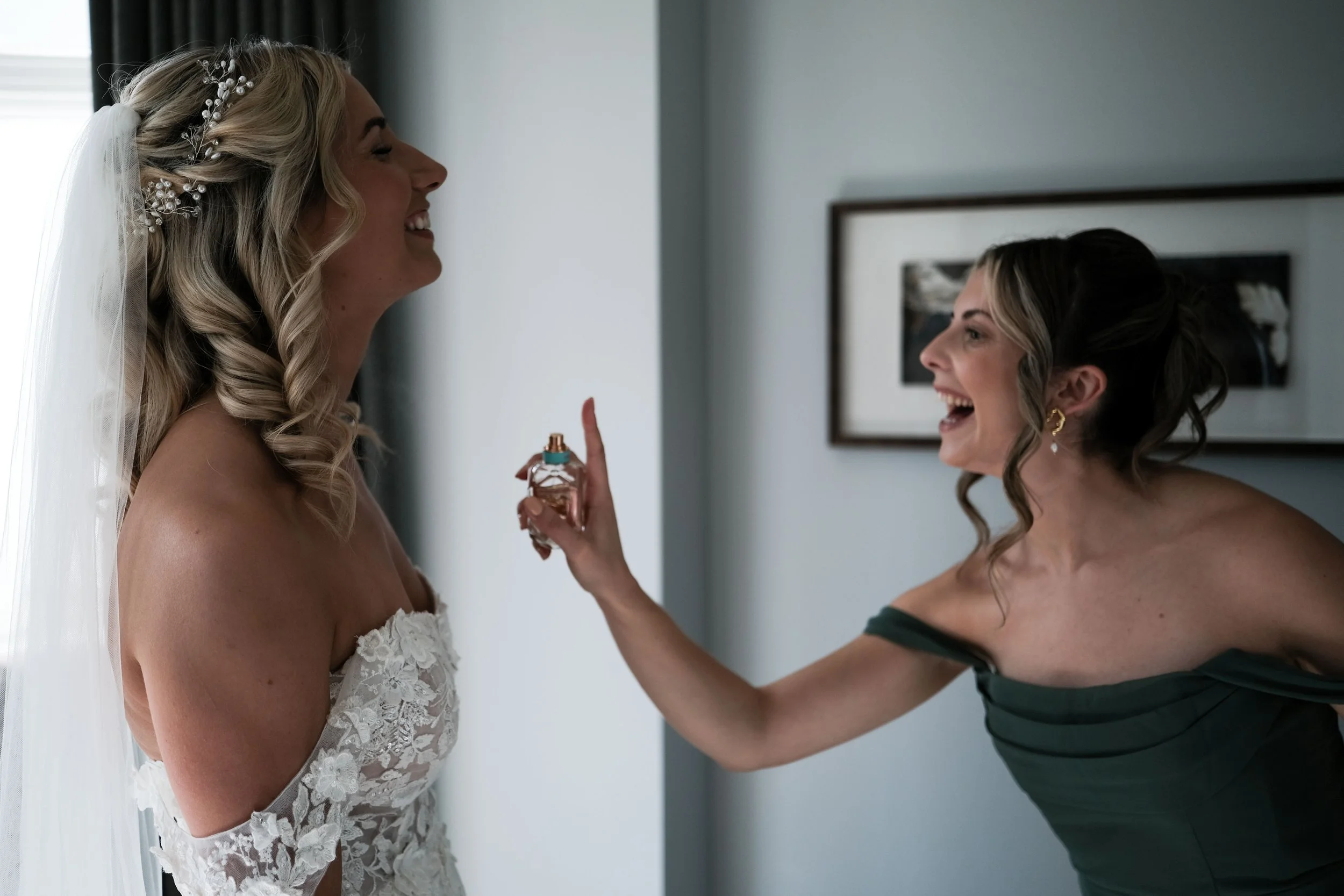 A bride in a lace wedding dress and veil smiling as another woman in a dark green off-shoulder dress playfully sprays perfume on her.