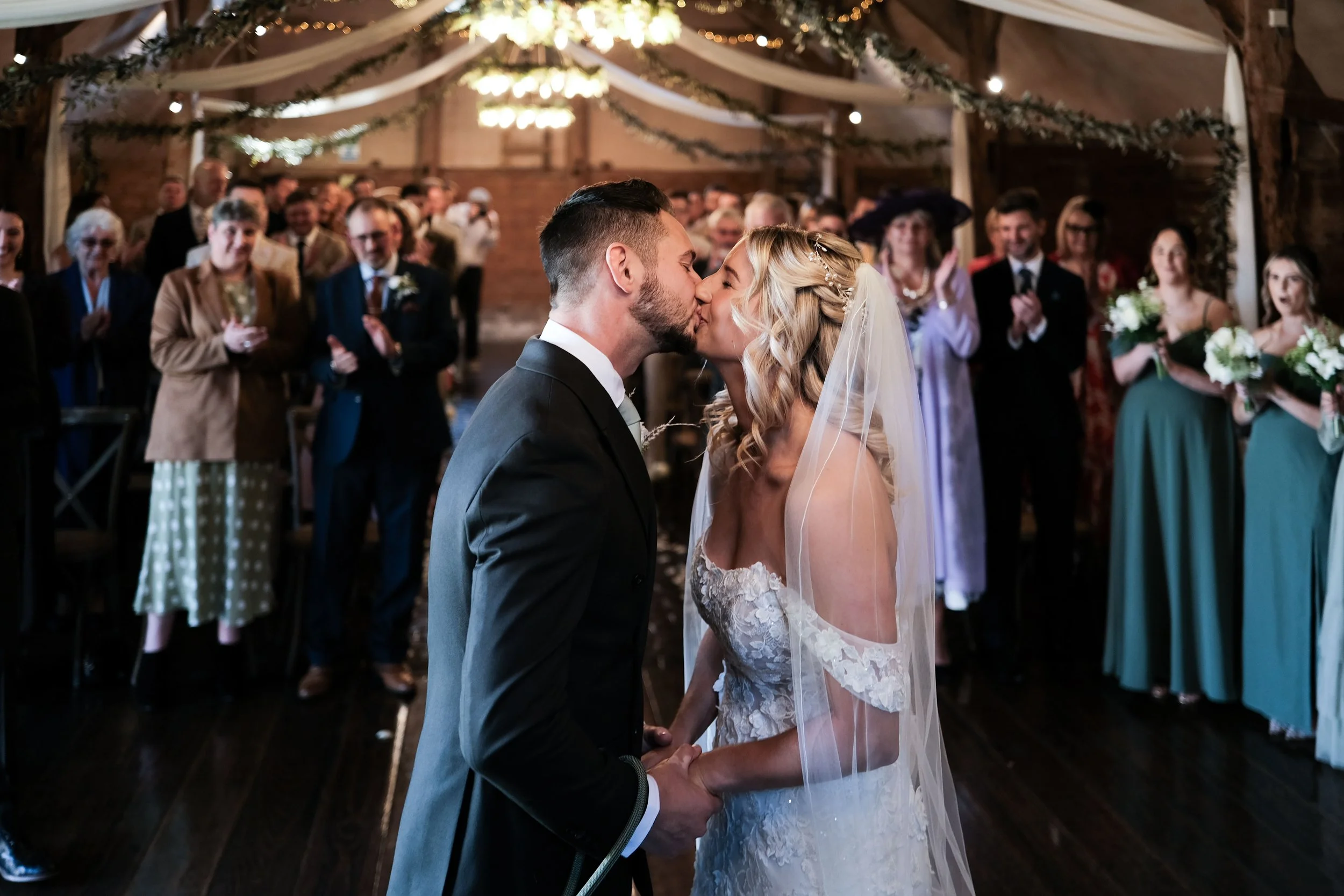 A bride and groom kiss during their wedding ceremony inside a decorated venue, with wedding guests clapping and watching in the background.
