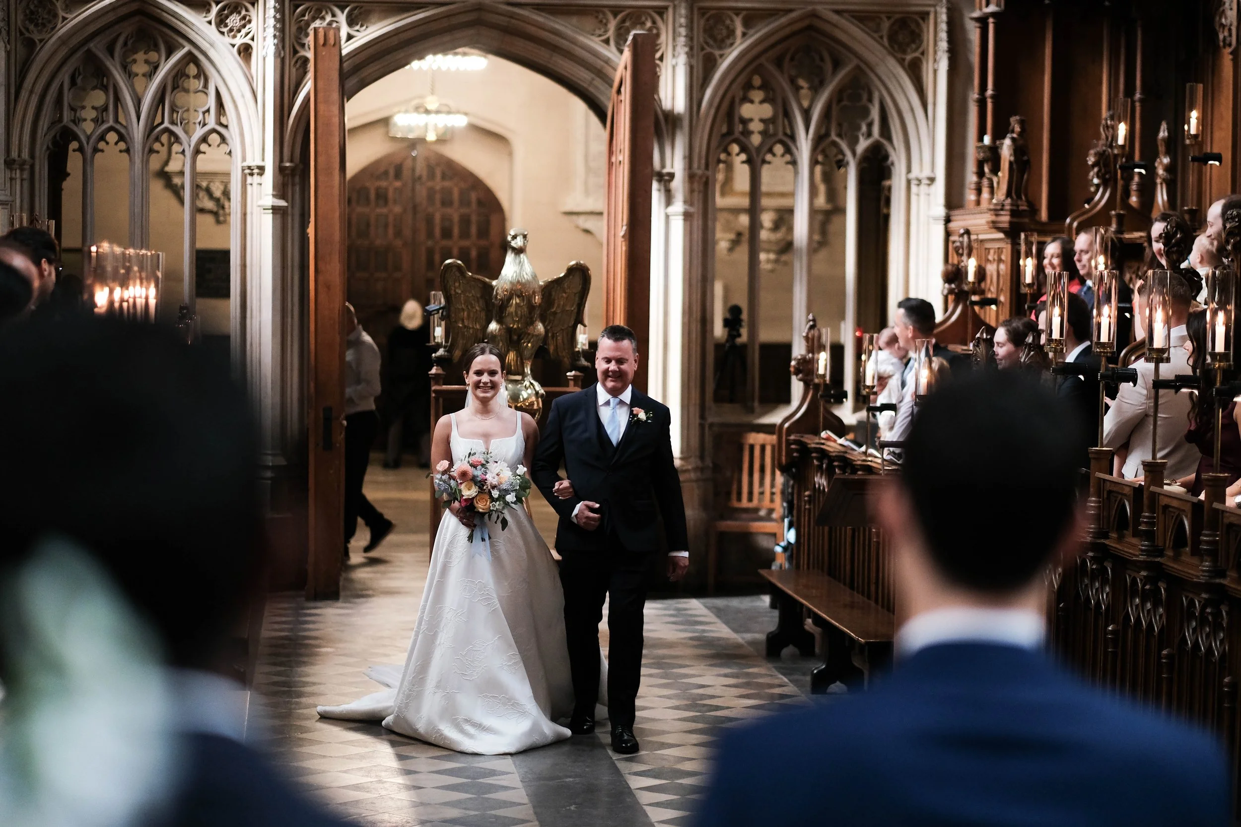 A bride walking down the aisle arm-in-arm with a man in a dark suit inside a church with Gothic architecture and candles